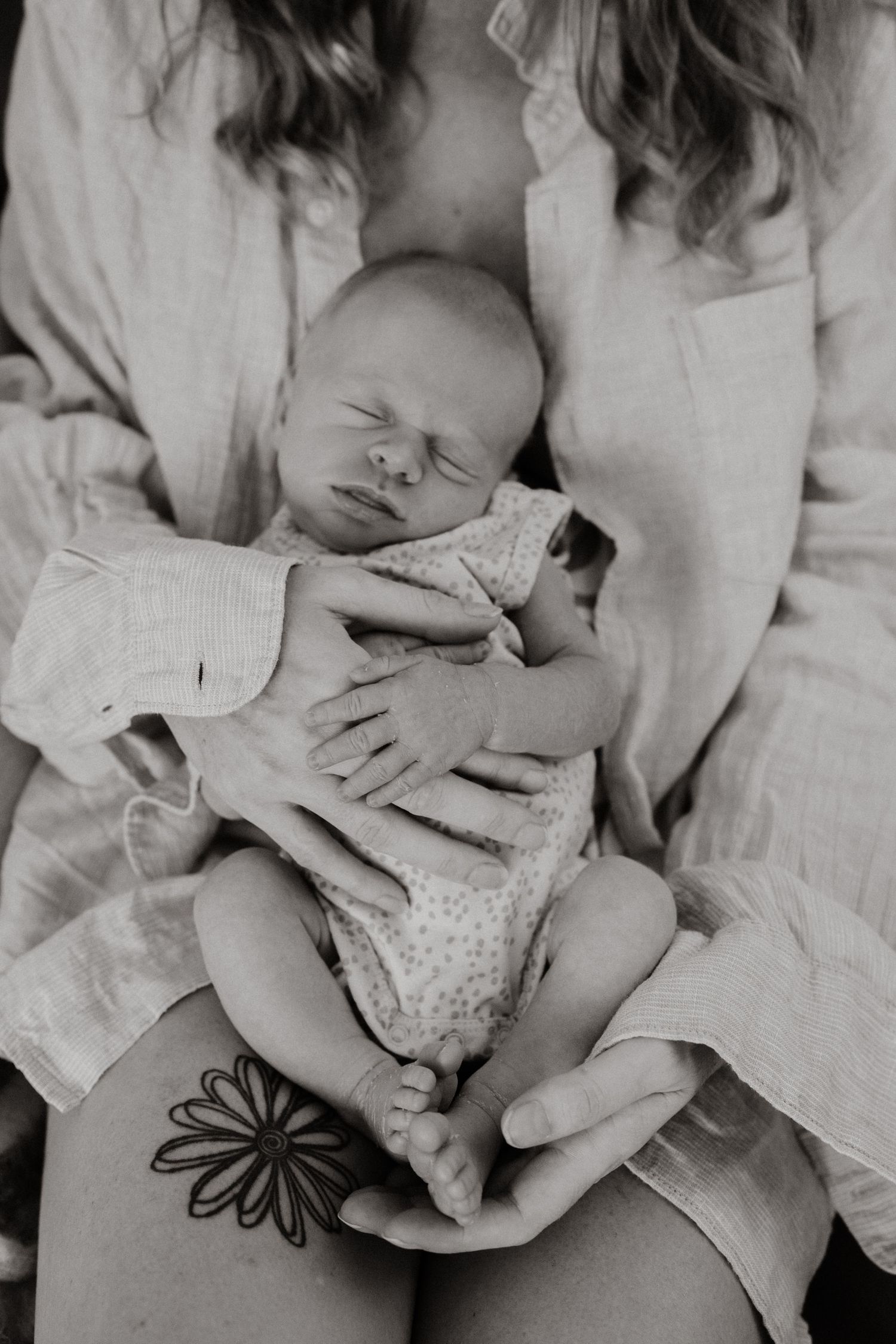 Black and white photo of mother holding sleeping newborn baby with floral tattoo visible on hand.