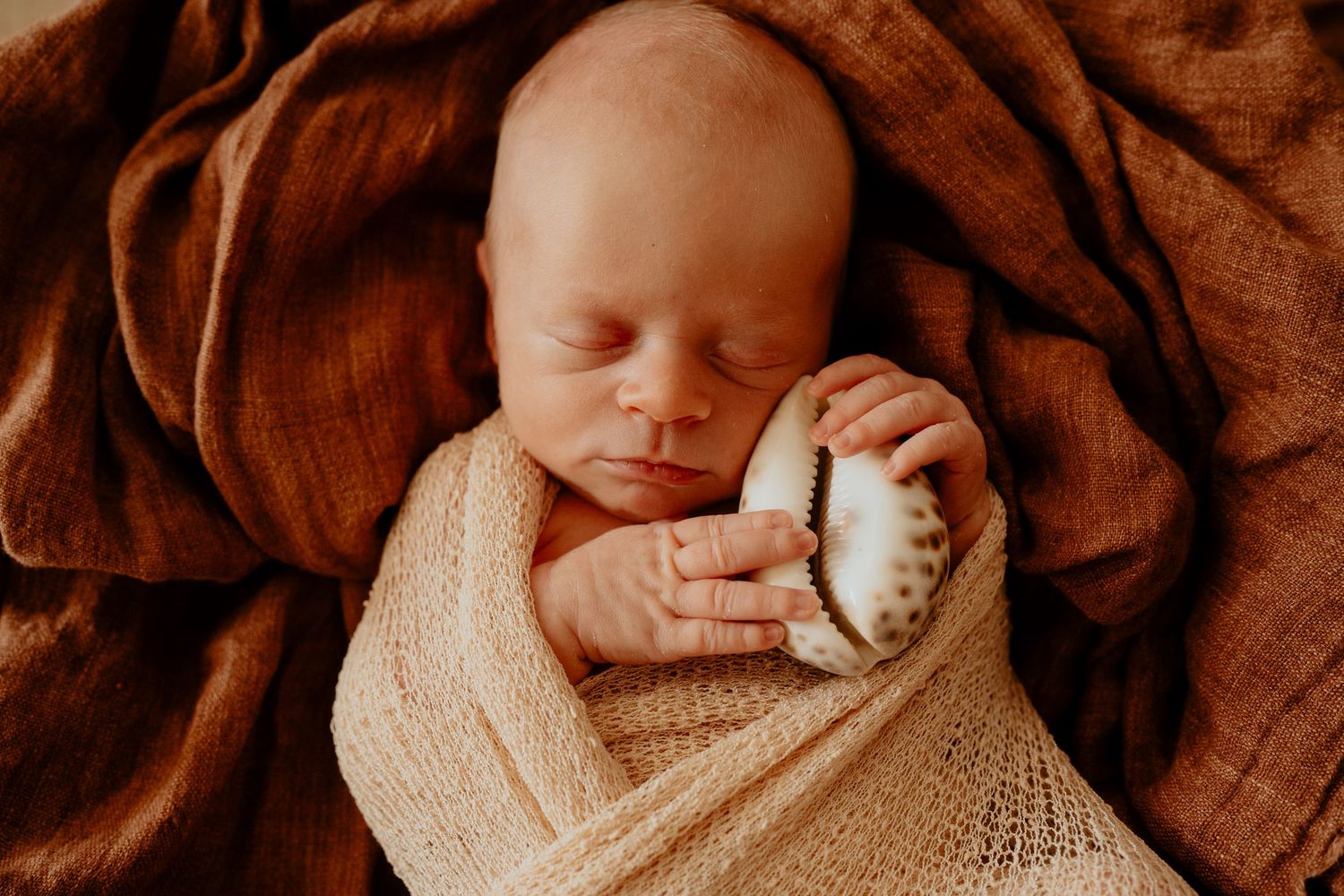 Peacefully sleeping newborn baby swaddled in a cream knit wrap holding a small shell.