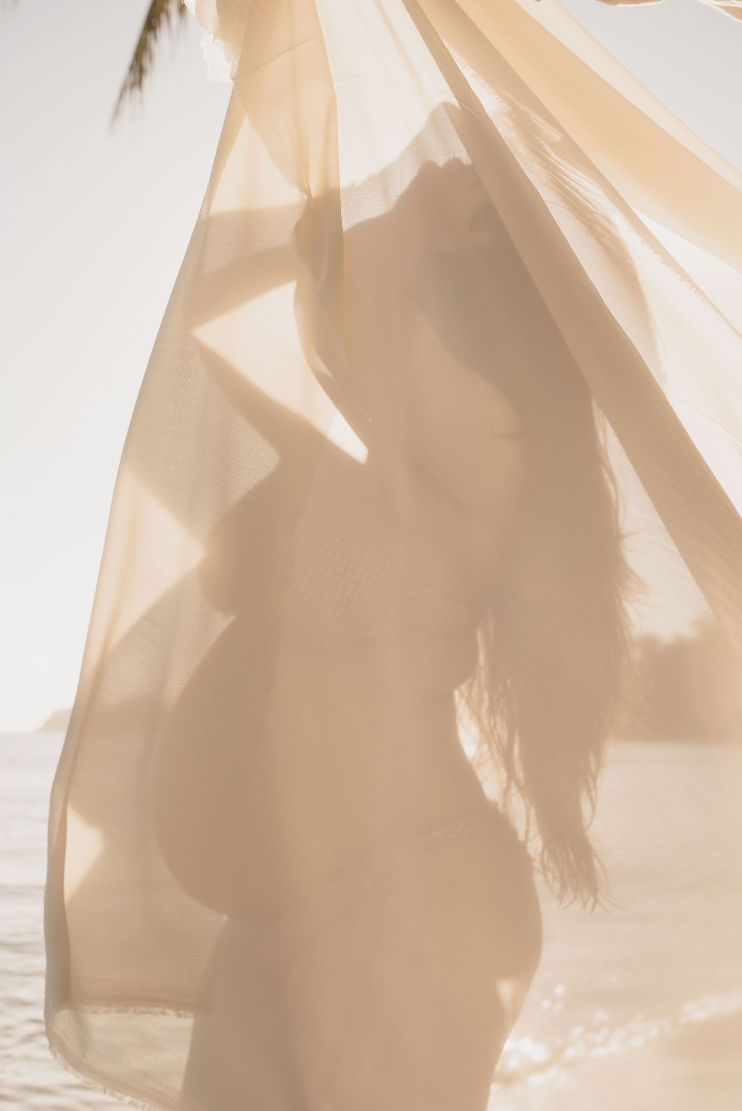 Artistic silhouette of pregnant woman draped in sheer flowing fabric against bright white sky.