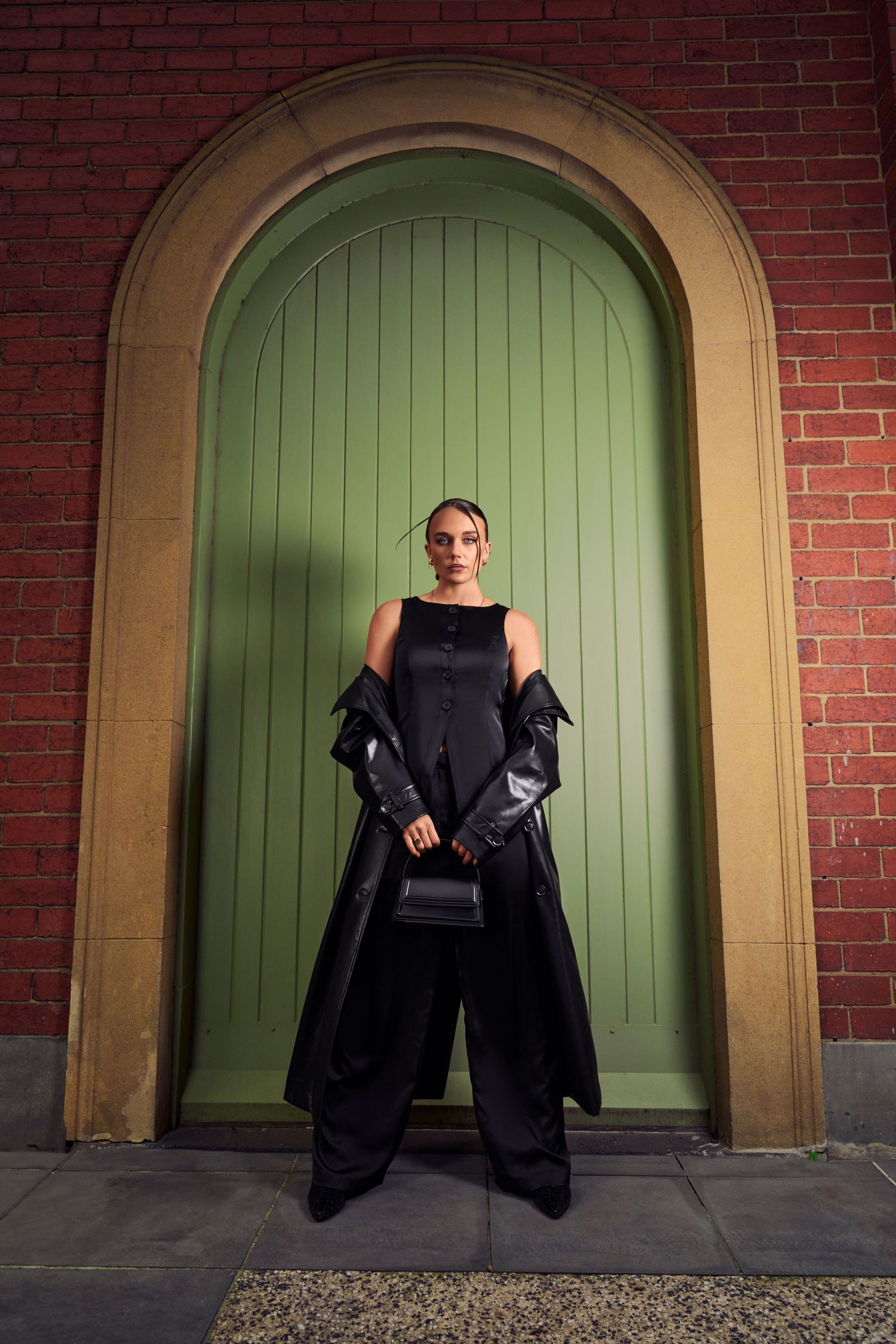 Woman in black wide-leg pants and sleeveless top with leather coat by green arched door.