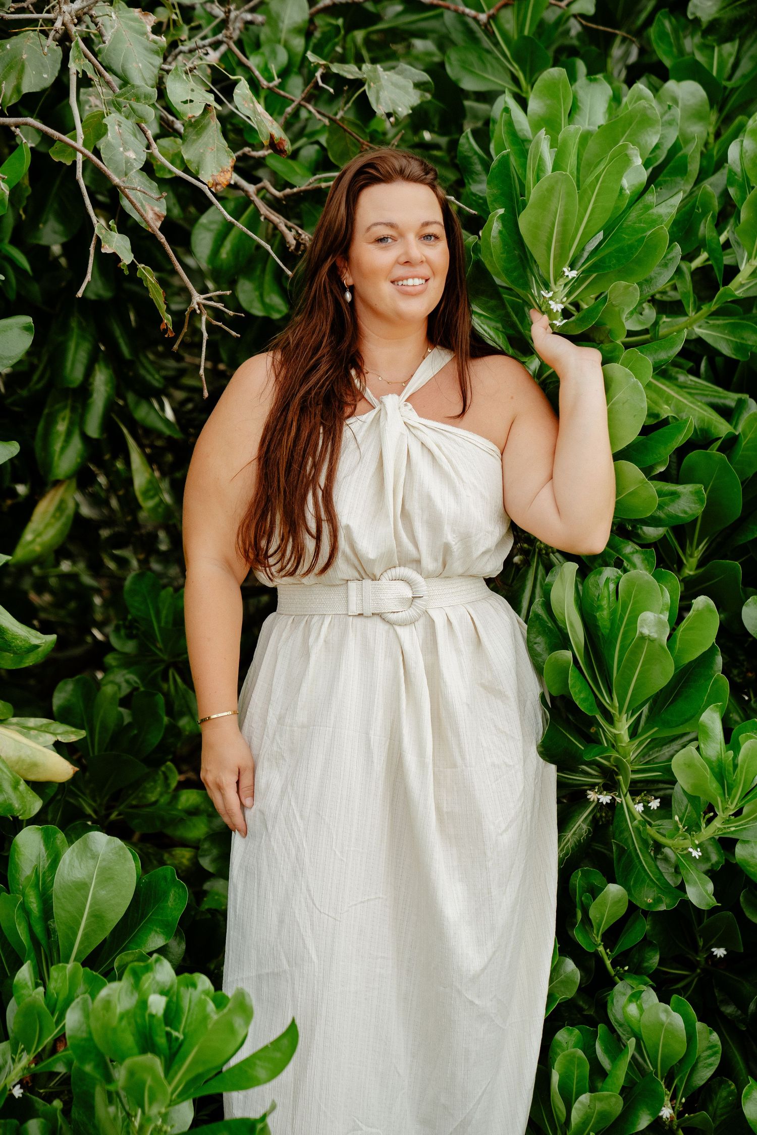 Smiling woman in white belted halter dress standing among lush green tropical foliage.