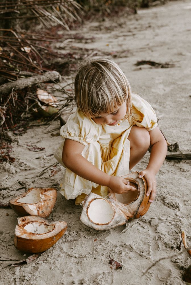 Young blonde girl in yellow dress examining a cracked open coconut on sandy beach.