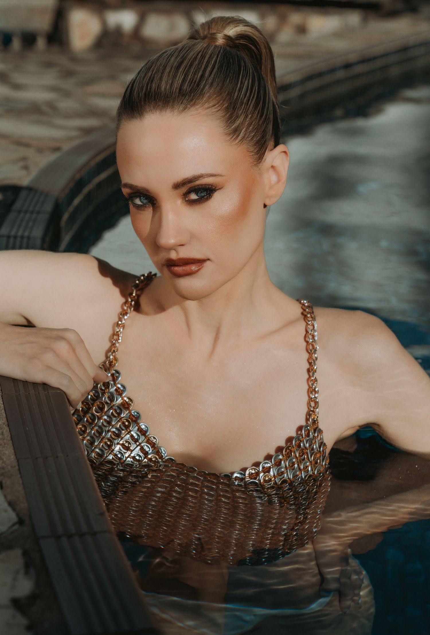 Elegant woman in a gold chainmail top posing in a swimming pool with a sultry expression.