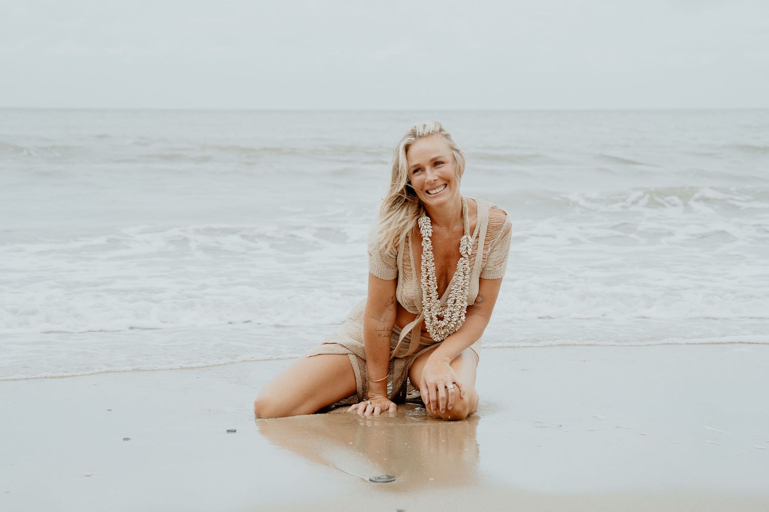 A smiling blonde woman in a beige dress kneels on a wet sandy beach with ocean waves behind her.