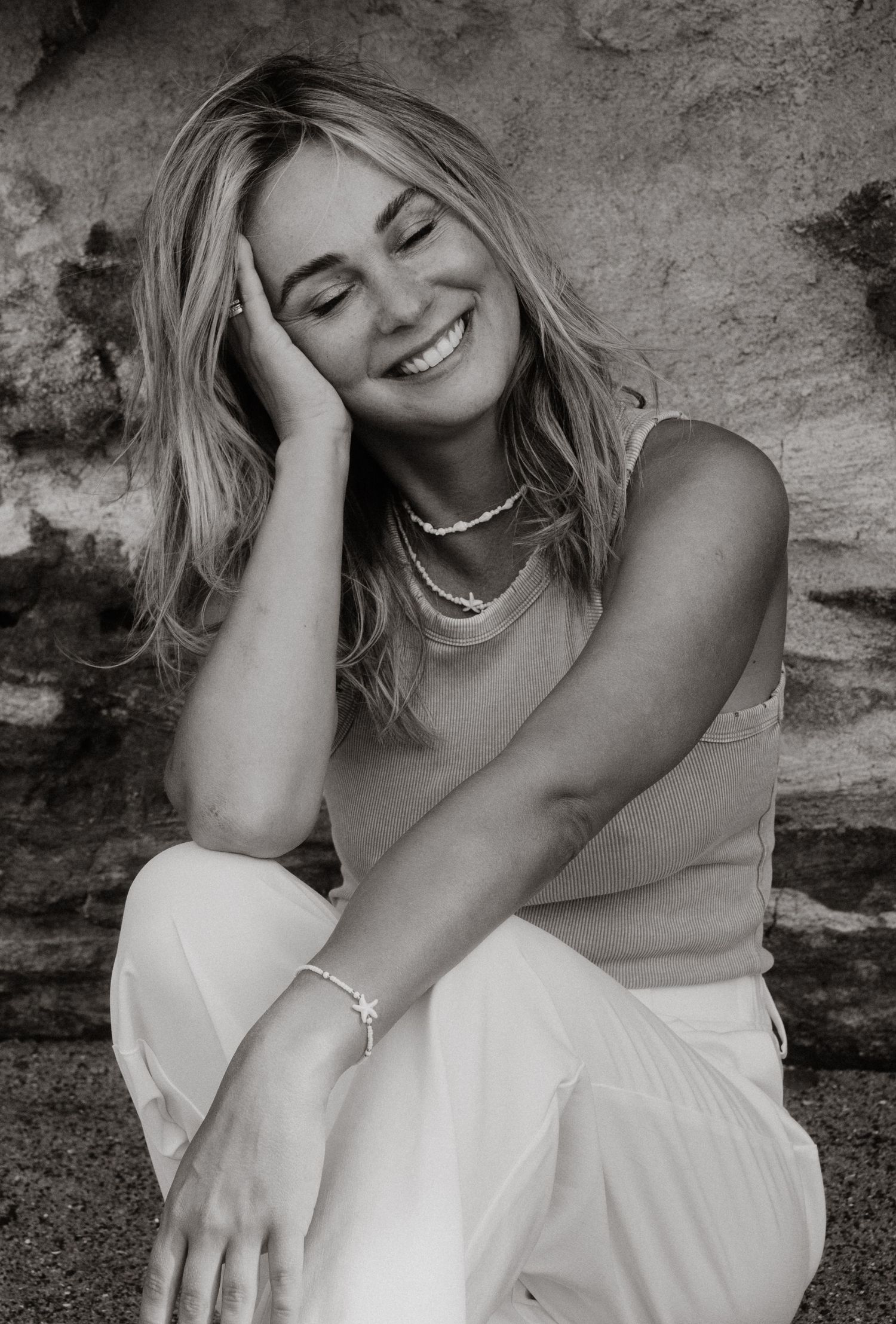 Black and white portrait of a smiling blonde woman sitting against a rock wall.