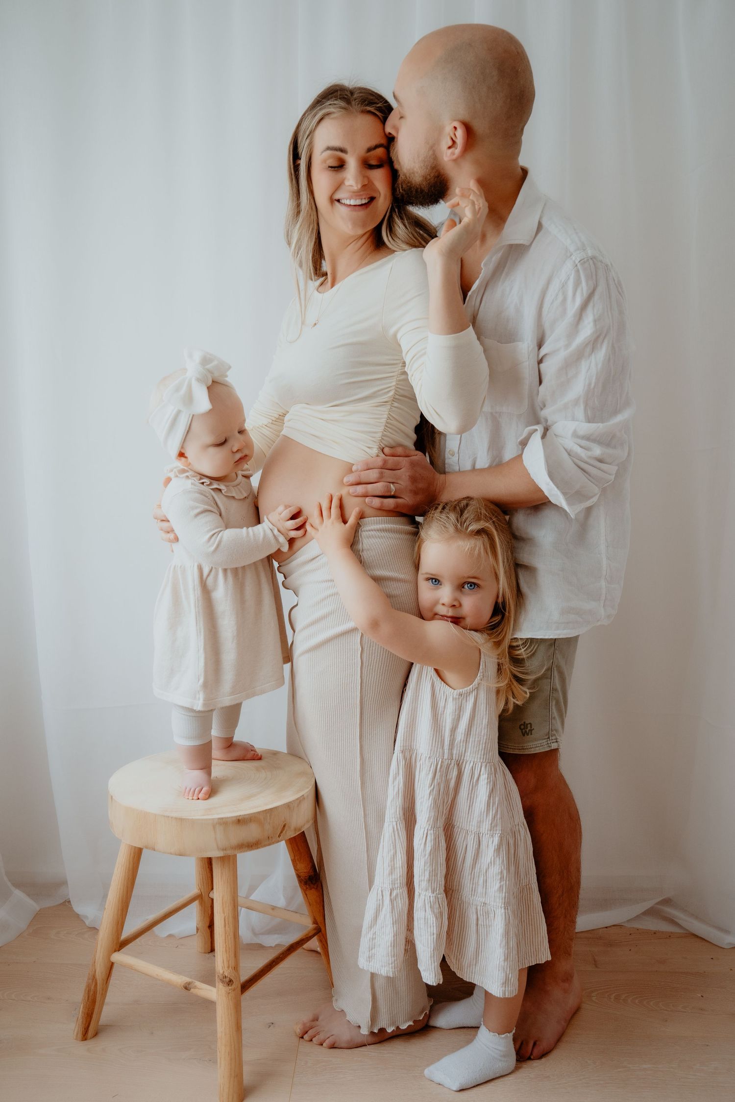 A happy family with a pregnant mother, bald father, and two young daughters pose together in neutral tones indoors.