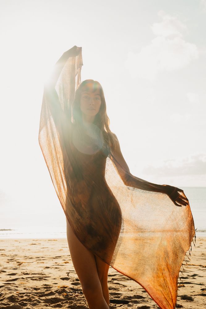 Woman holding sheer orange scarf on sunny beach with ocean backdrop.
