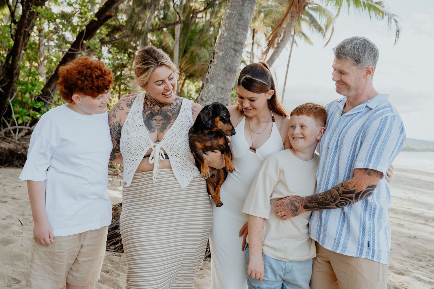 Blended family with dog smiles together on palm-lined beach in casual outfits.