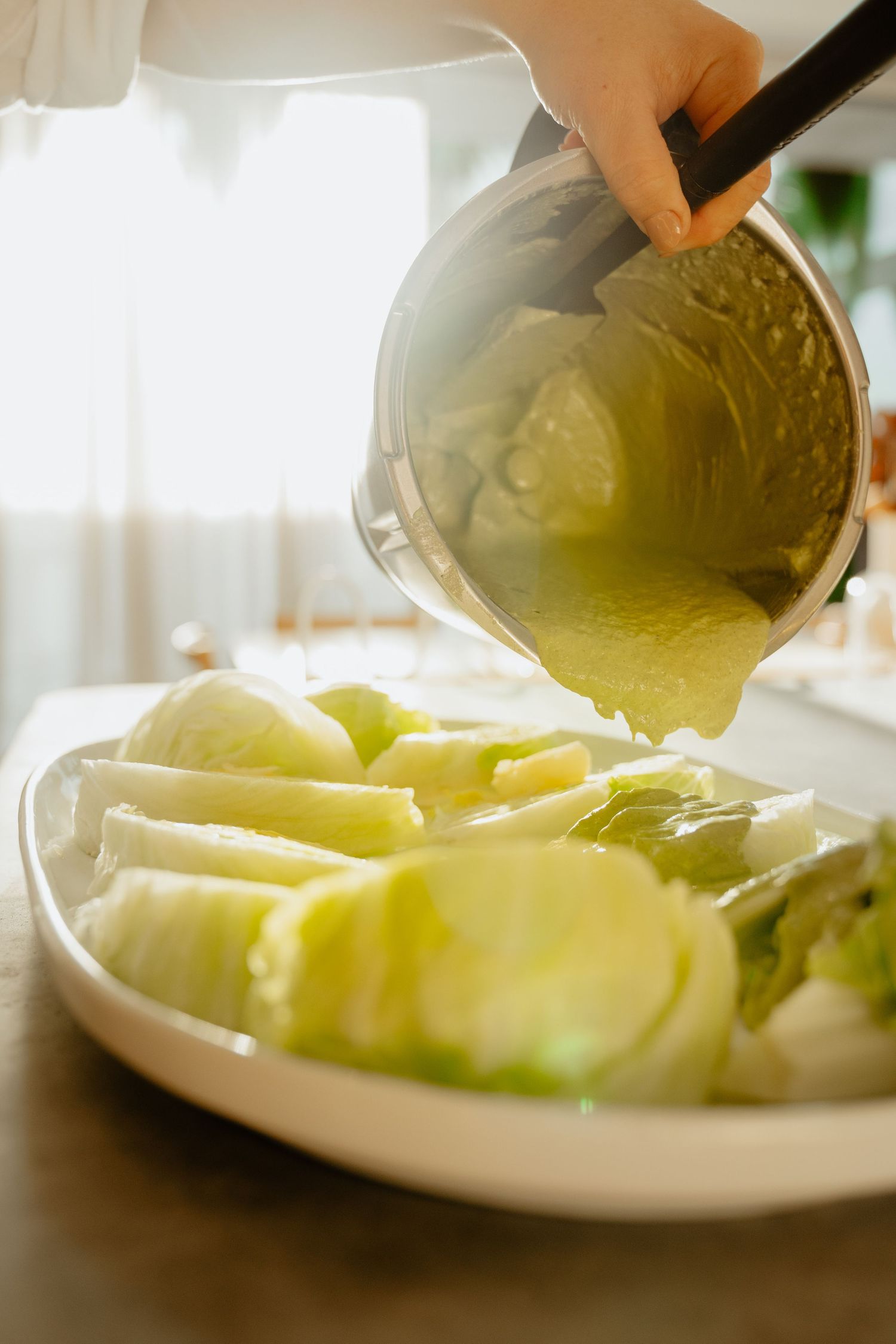 Person pouring cooked cabbage from a pot onto a white serving plate in a sunlit kitchen.