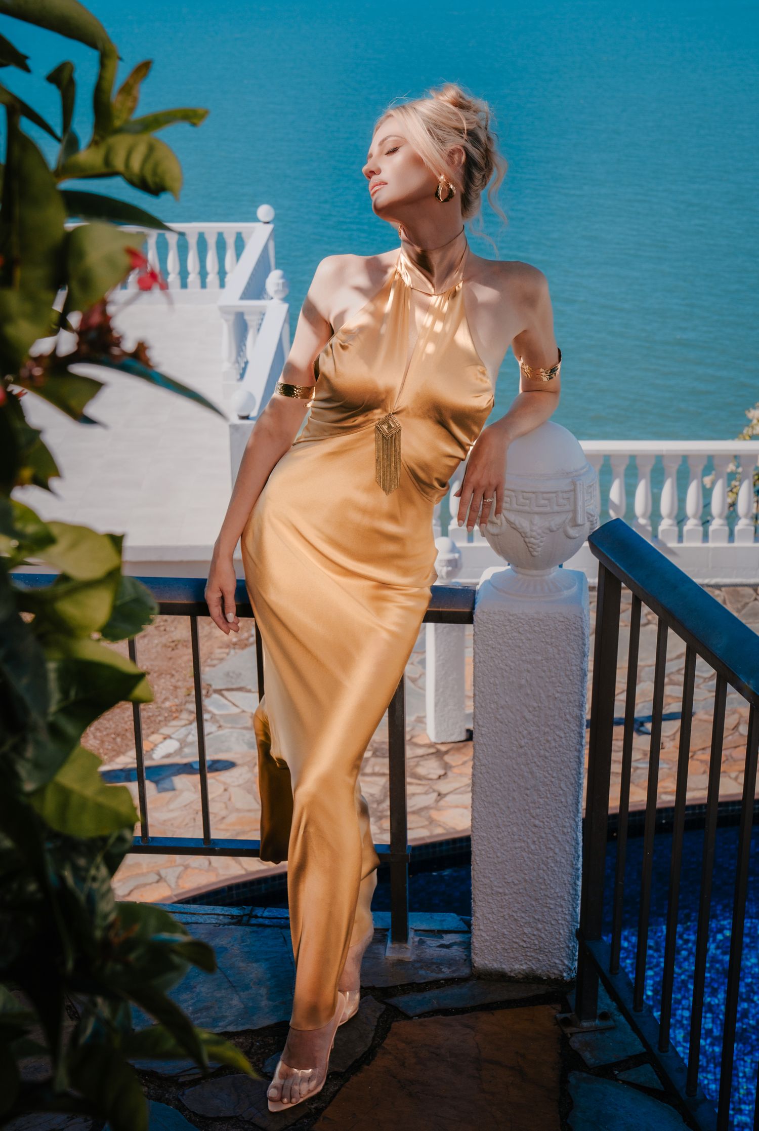 Elegant blonde woman in a gold satin halter maxi dress posing on a sunlit Mediterranean balcony.