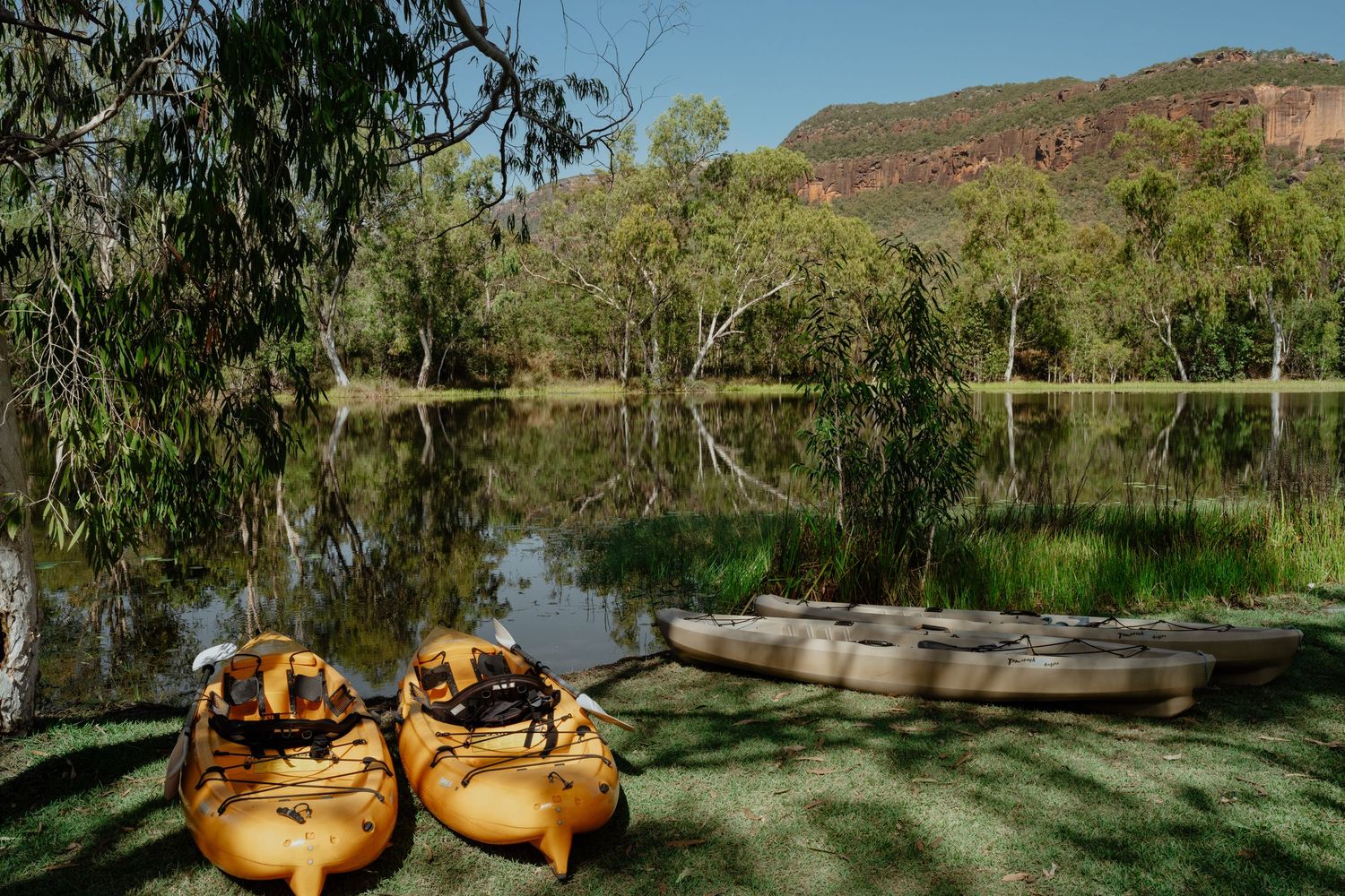 Two yellow kayaks resting on green grass beside a calm river with red rocky mountains in background.