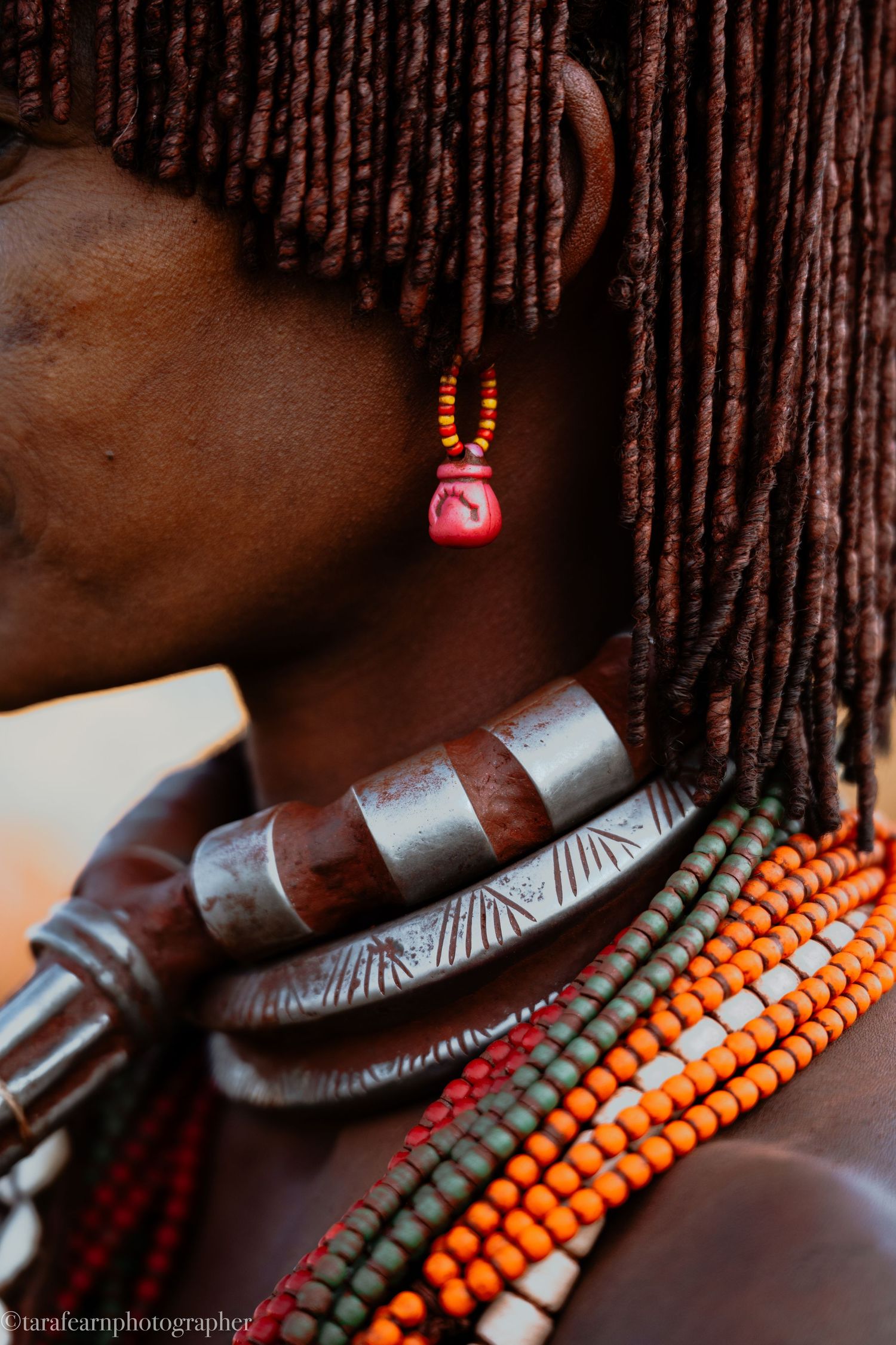 Close-up of African woman wearing traditional metal collar necklace, colorful beads, and red earring with braided hair.