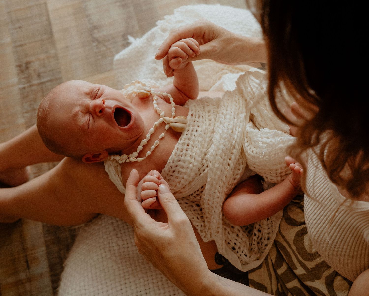 Yawning newborn baby in white crochet wrap being held by mother with pearl necklace accessory.