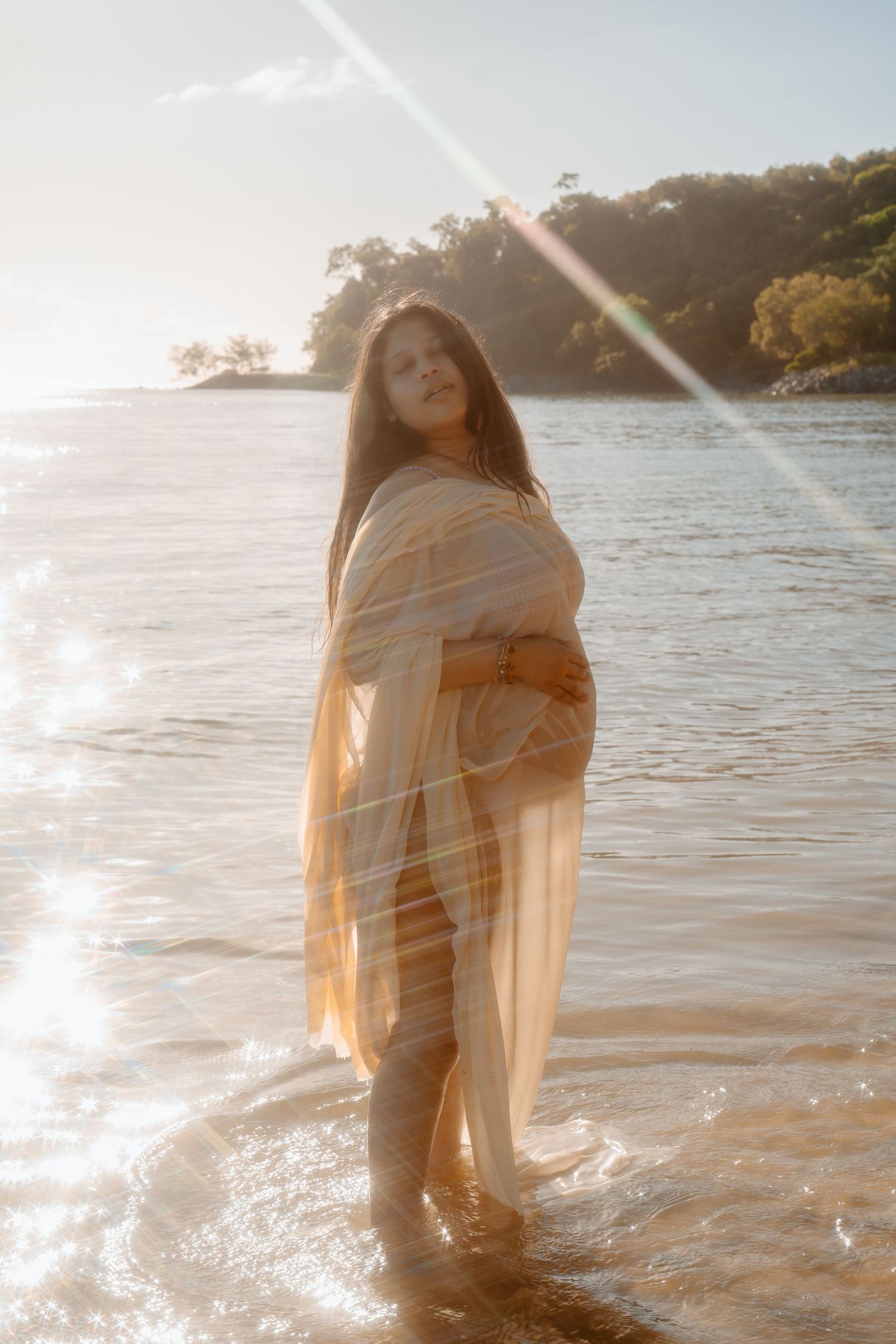 Pregnant woman wrapped in sheer fabric stands in shallow water at golden hour beach.