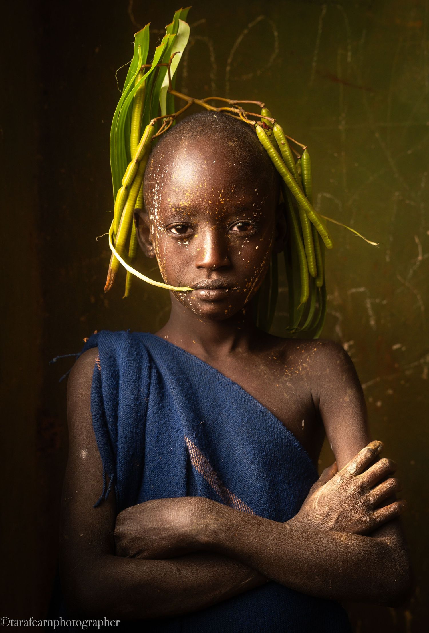 Young African child with green leaves as a headdress, wrapped in blue cloth, arms crossed, dramatic lighting.