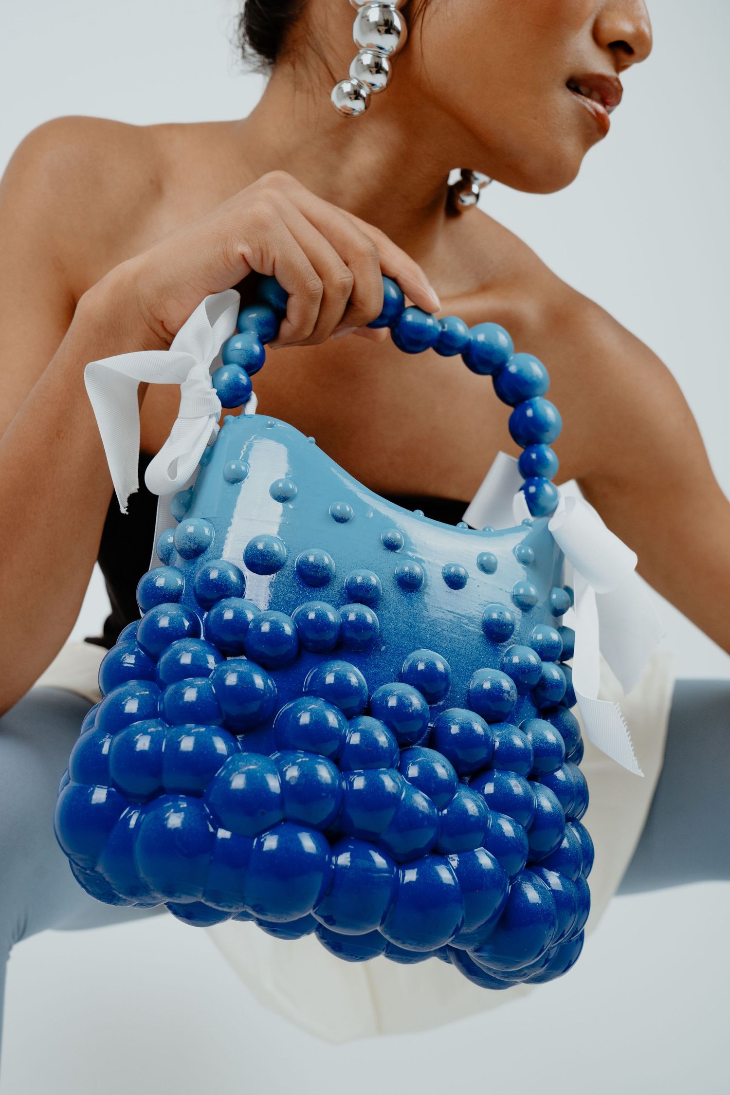 Woman holding a unique blue bubble-textured handbag with beaded handle and silver ball earrings.