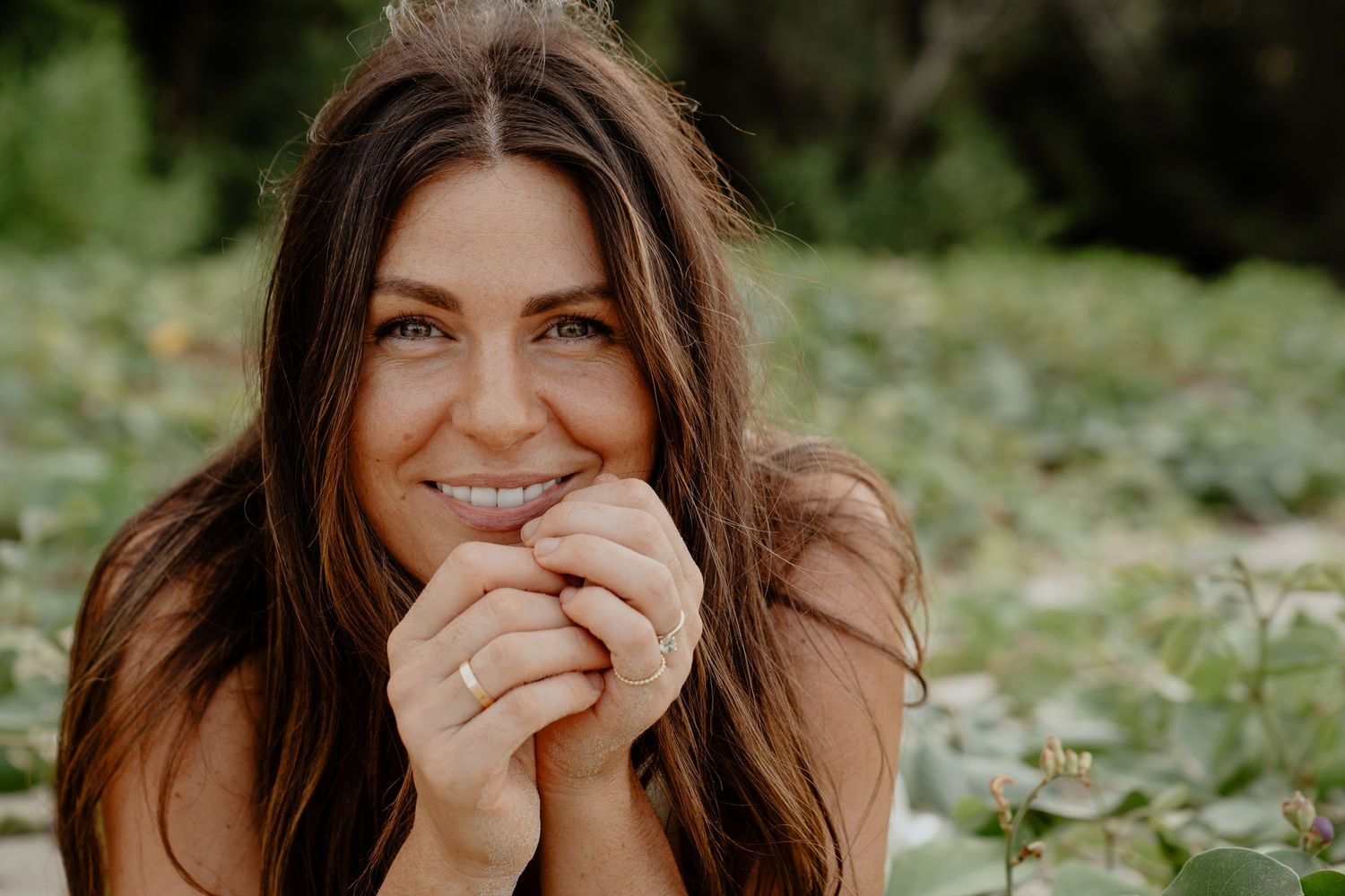 Close-up of smiling brunette woman with hands clasped surrounded by green foliage.