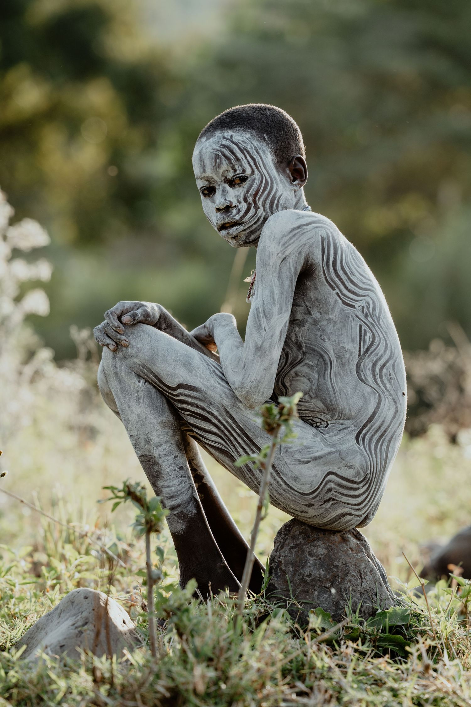 Child with grey clay body paint sitting on a rock in a natural outdoor setting.