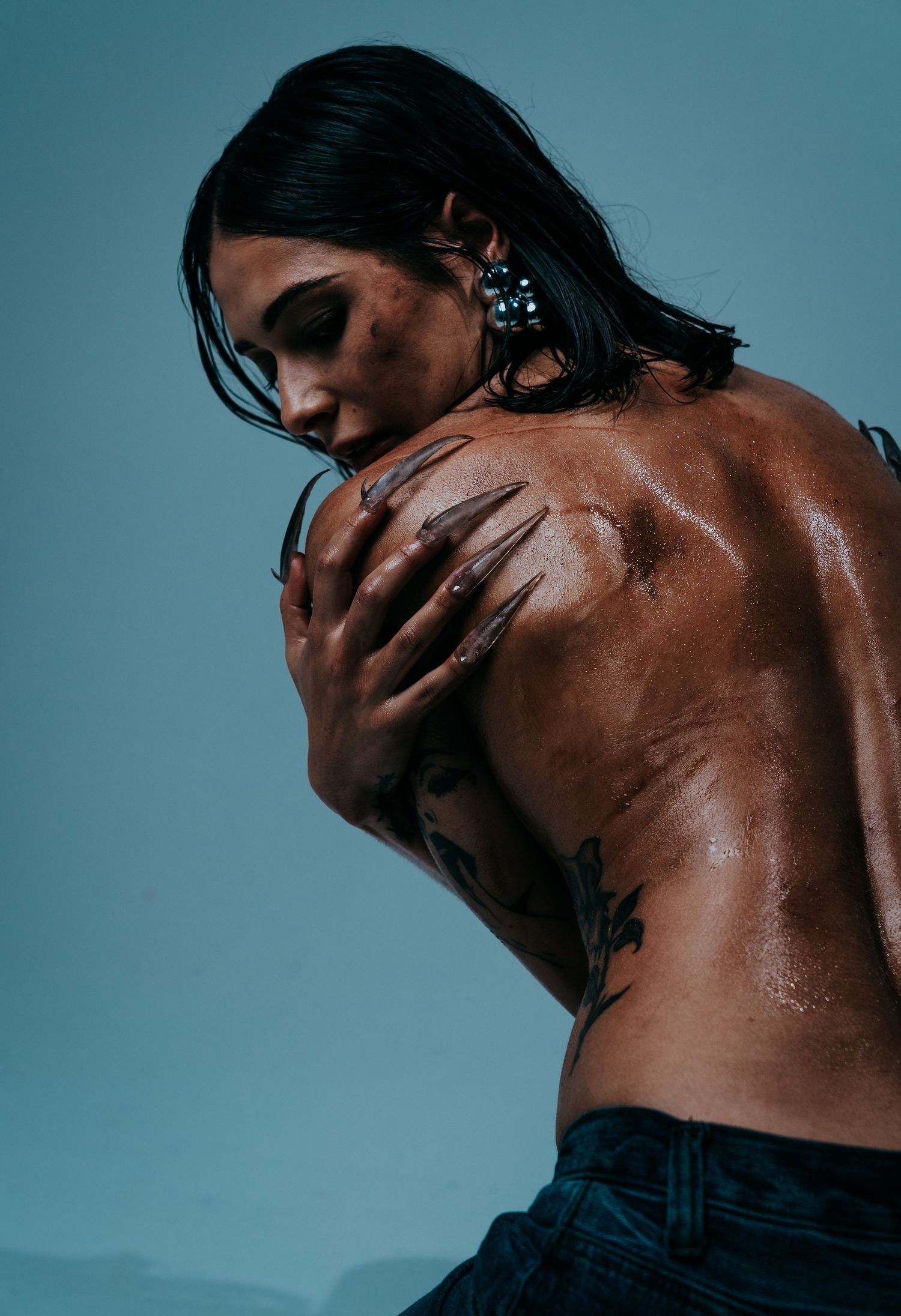 Woman with back tattoos posed with wet skin against blue-grey studio background.