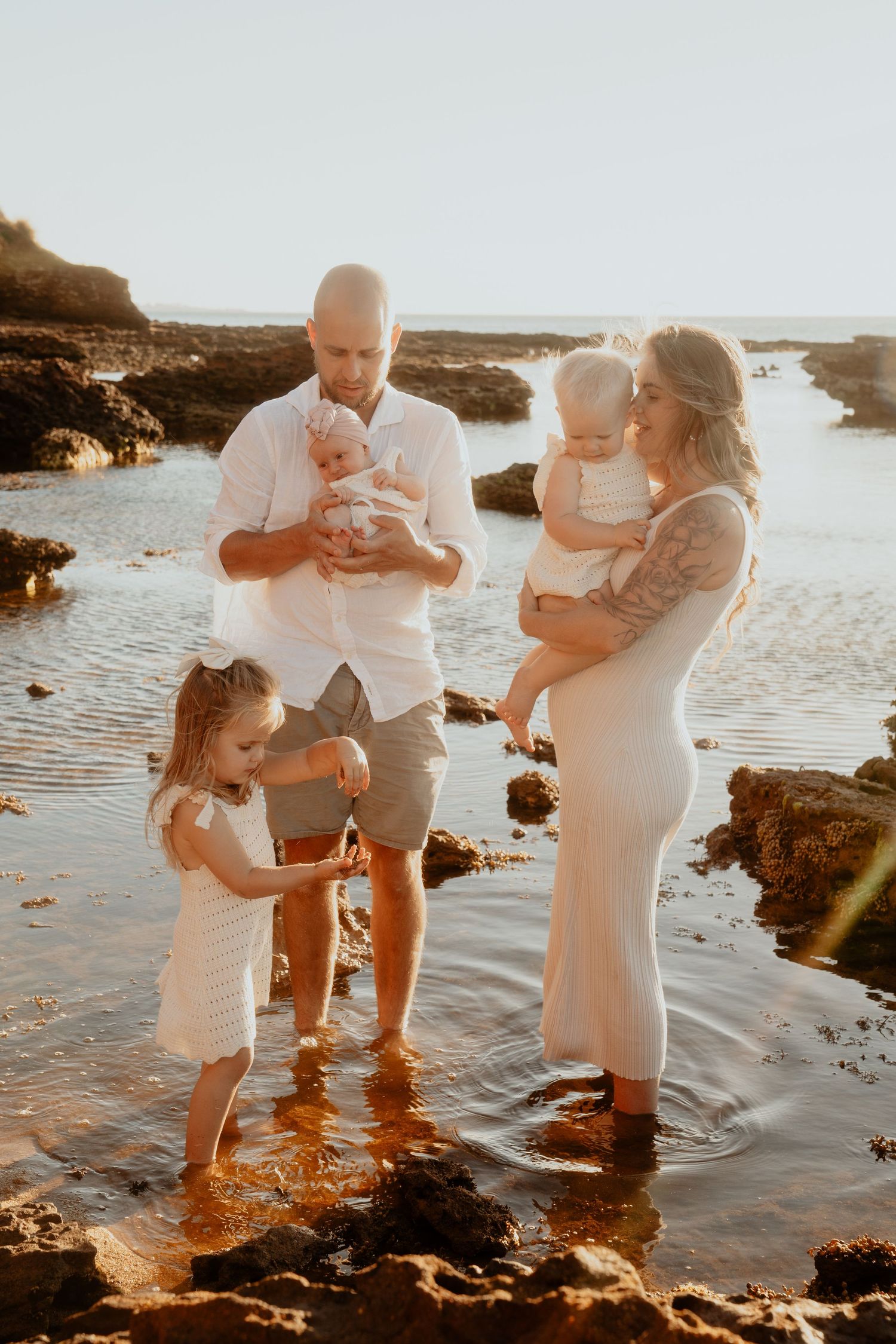 Happy family of five standing in shallow coastal water at sunset, parents holding two babies.