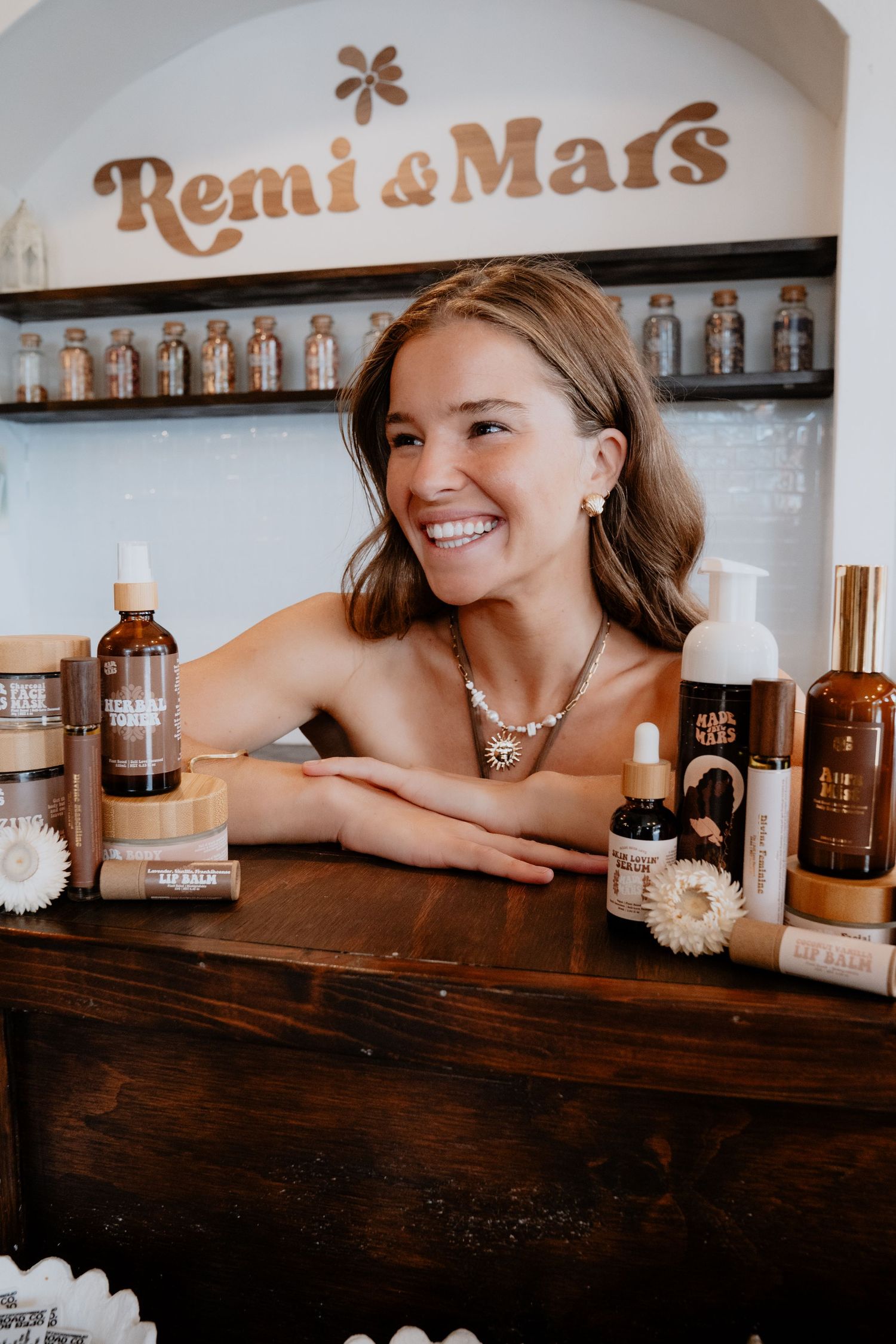 Smiling woman leaning on wooden counter surrounded by Remi and Mars skincare products.