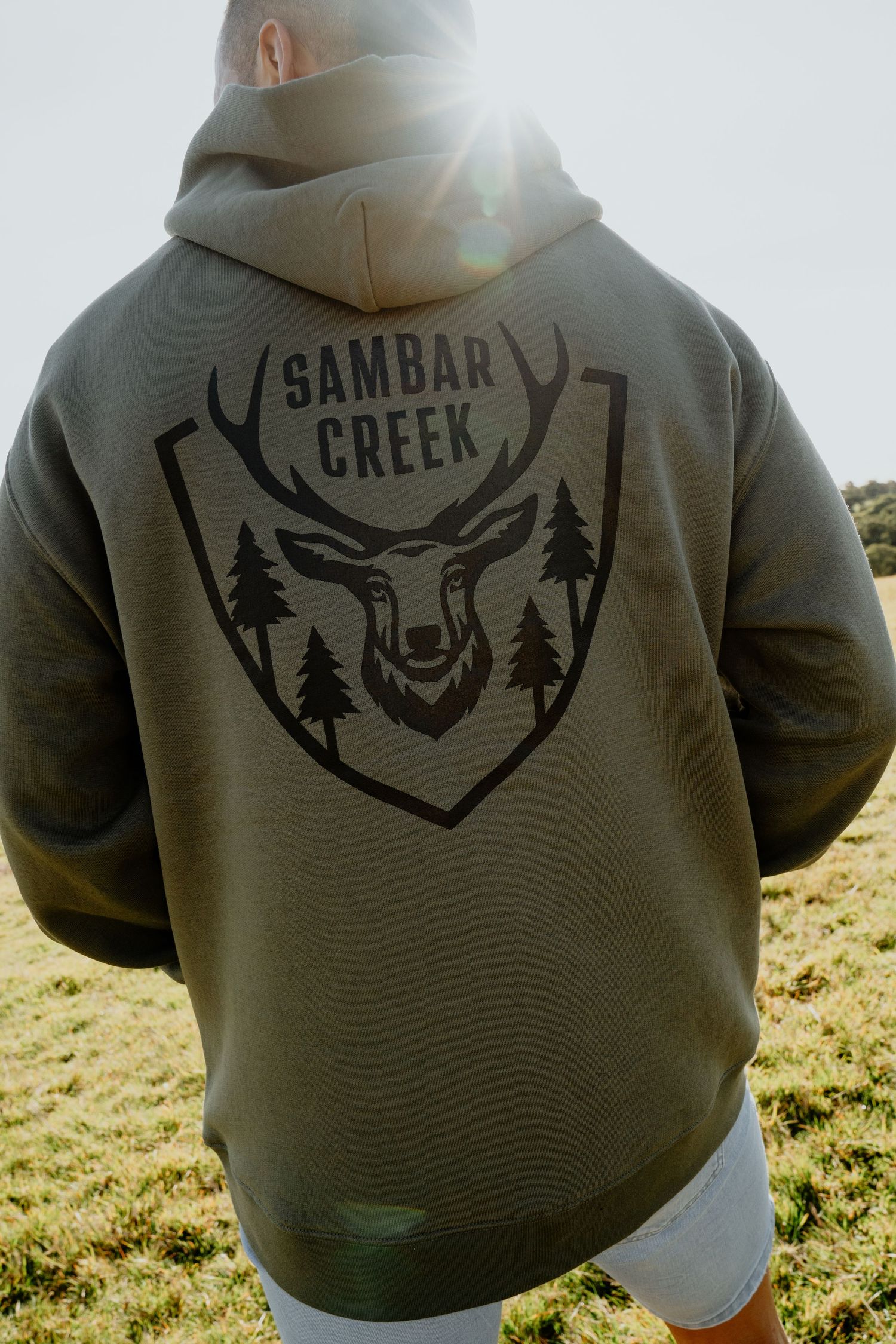 Man wearing olive green Sambar Creek hoodie with deer logo graphic on back, outdoors.