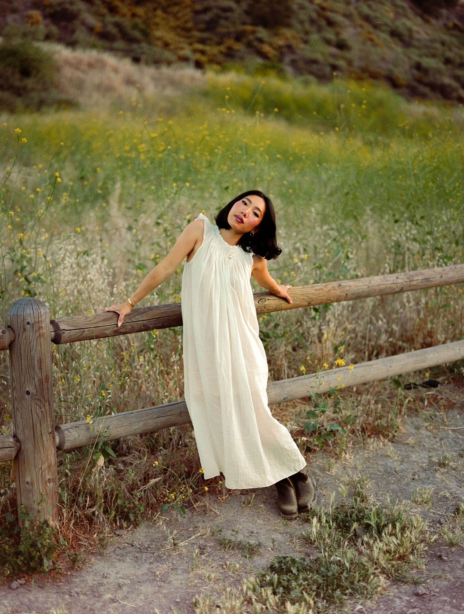 A girl in a white dress plays along a rustic wooden fence in a field of wildflowers in Los Angeles, CA.
