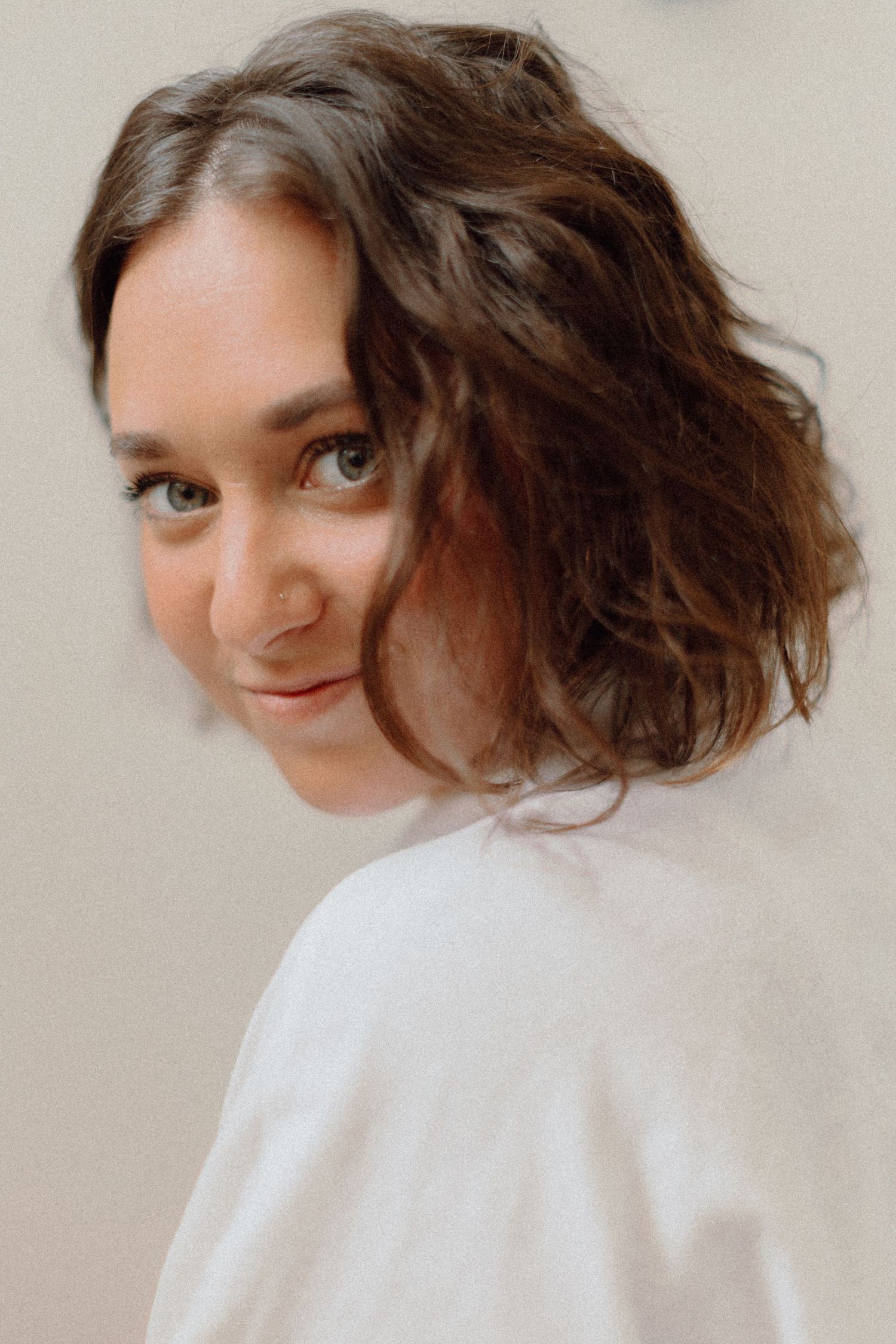 A portrait of someone with wavy brown hair and bright eyes looking over their shoulder in a white top against a neutral background.