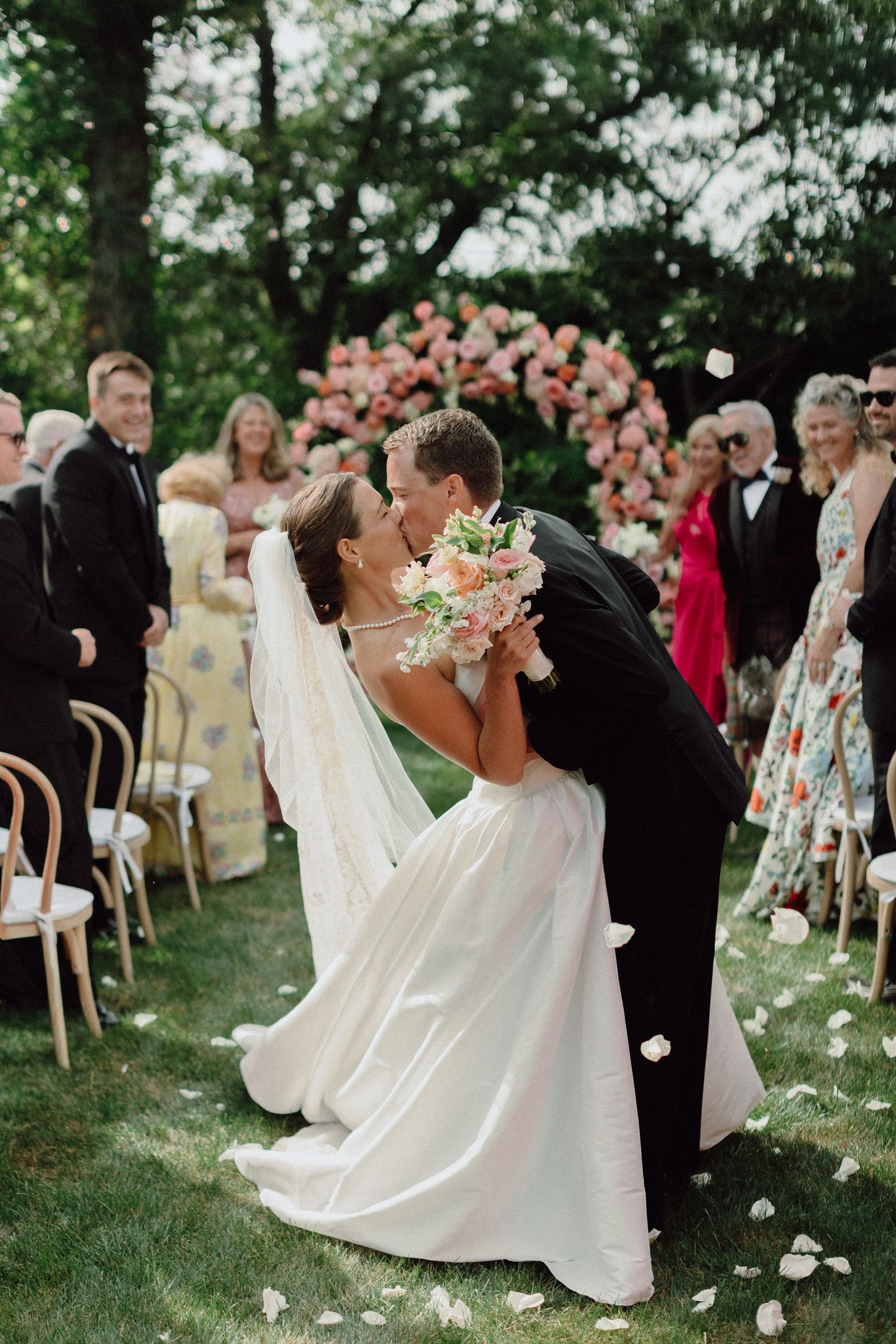 A romantic dip kiss between newlyweds during an outdoor garden wedding ceremony with guests and floral decorations.