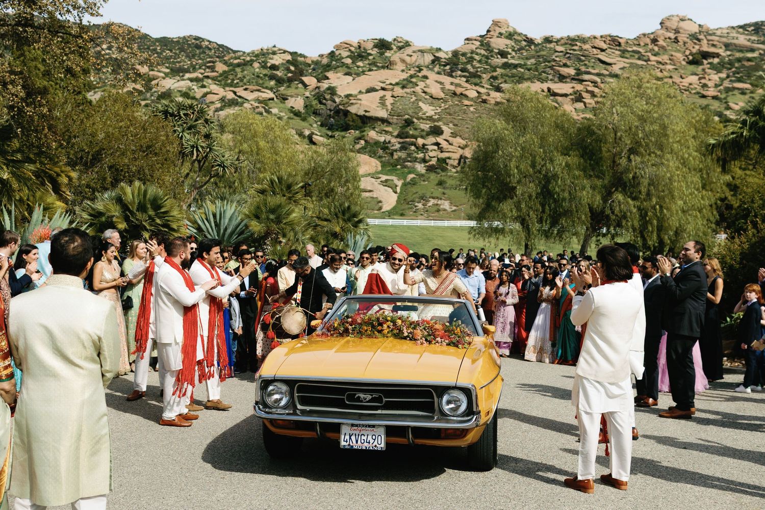 Indian wedding couple exits in vintage gold Ford Mustang surrounded by colorful guests in scenic outdoor venue.