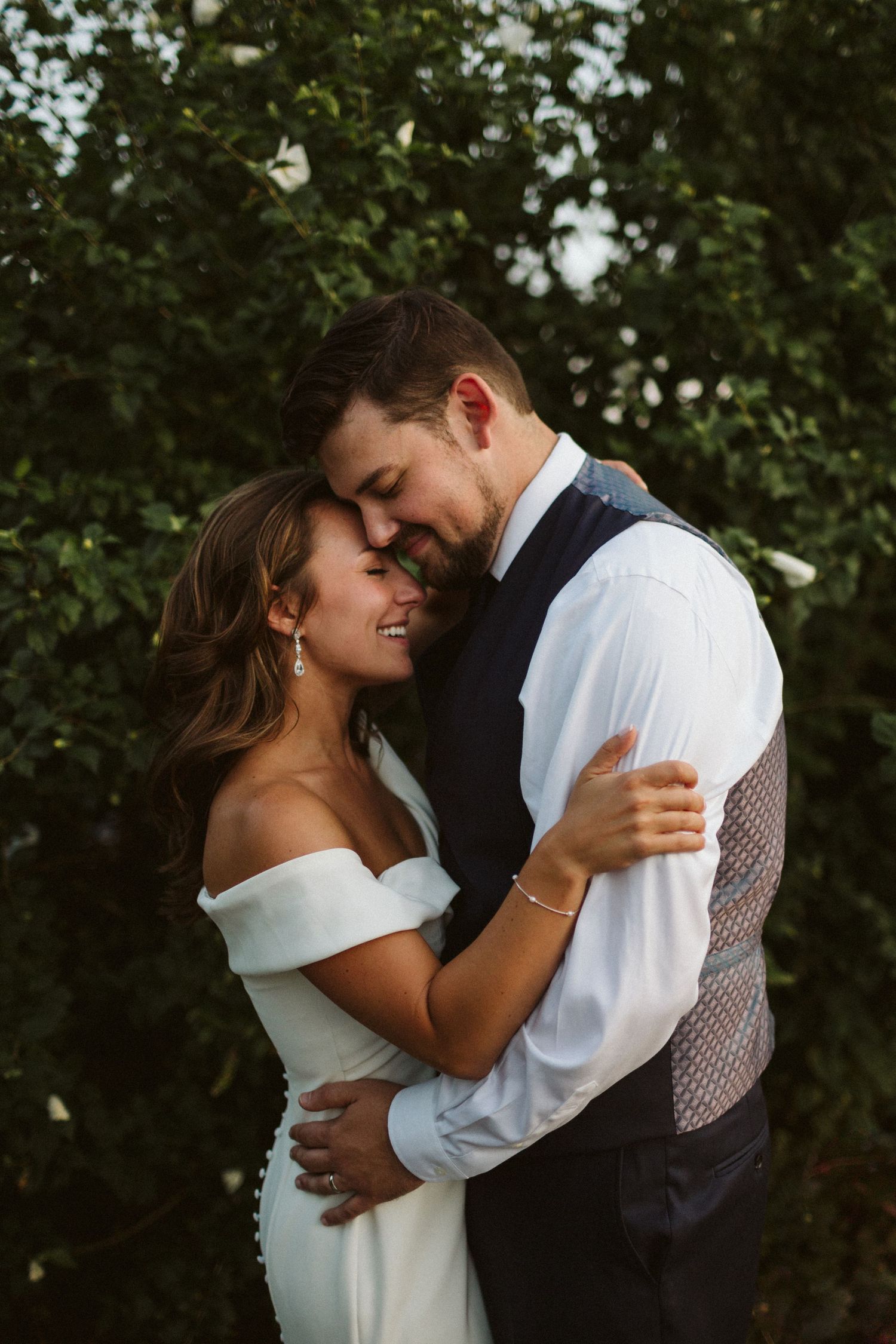 A romantic couple embraces intimately in a garden setting with lush greenery in the background.