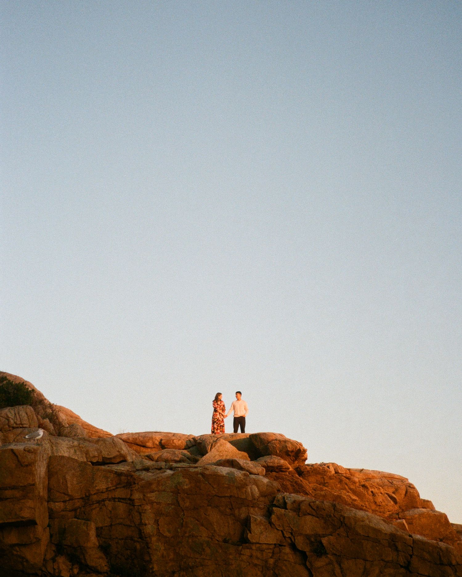 Two people standing on red rocks against a clear blue sky in Beverly, MA.