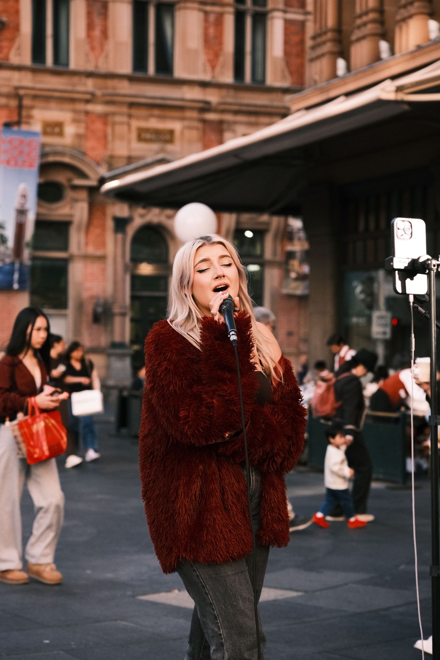 Young blonde woman singing into microphone outdoors, wearing red fur coat, busking on city street.