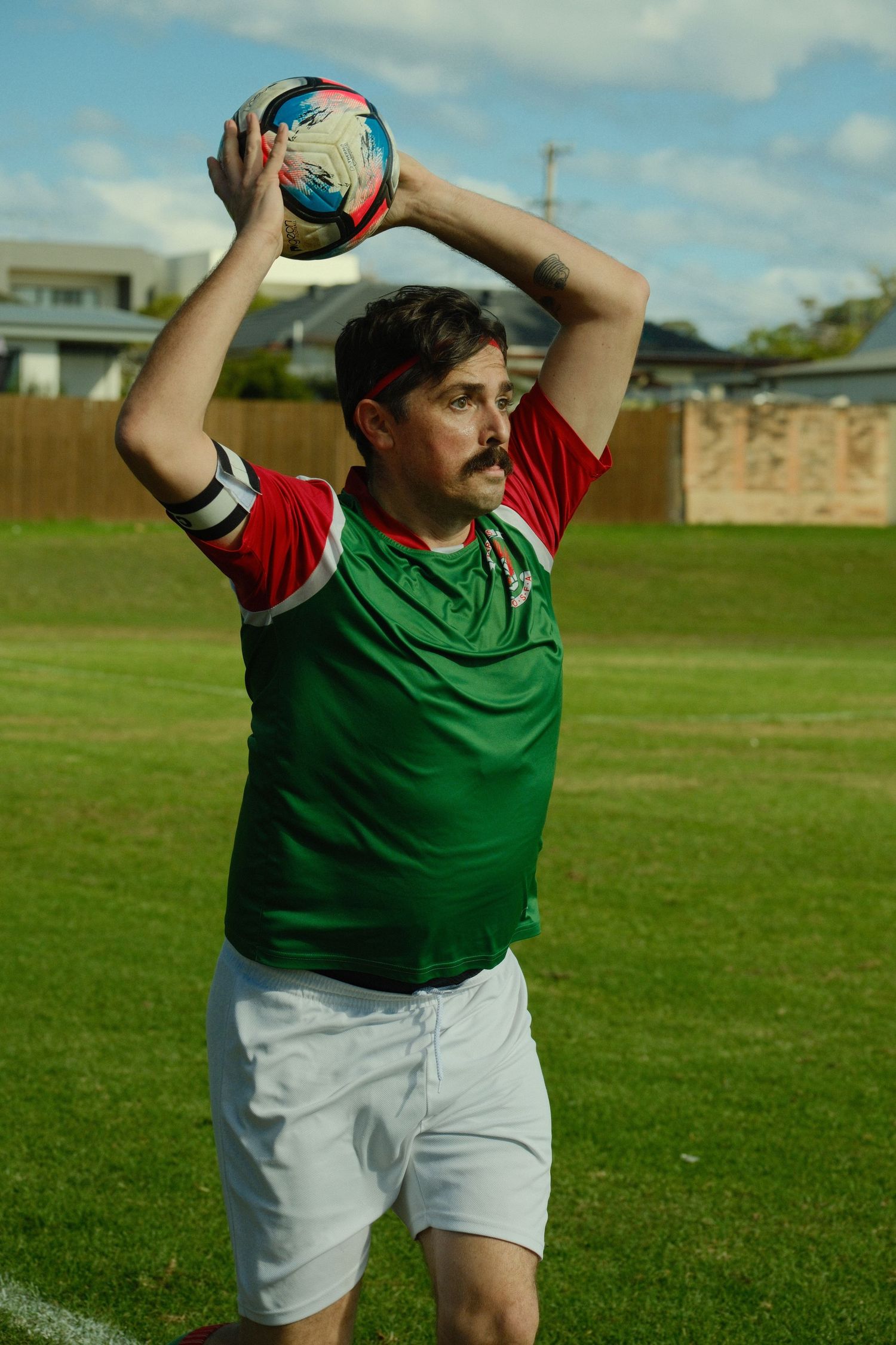 Man in green and red soccer jersey performs throw-in on grass field, holding ball overhead with both hands.