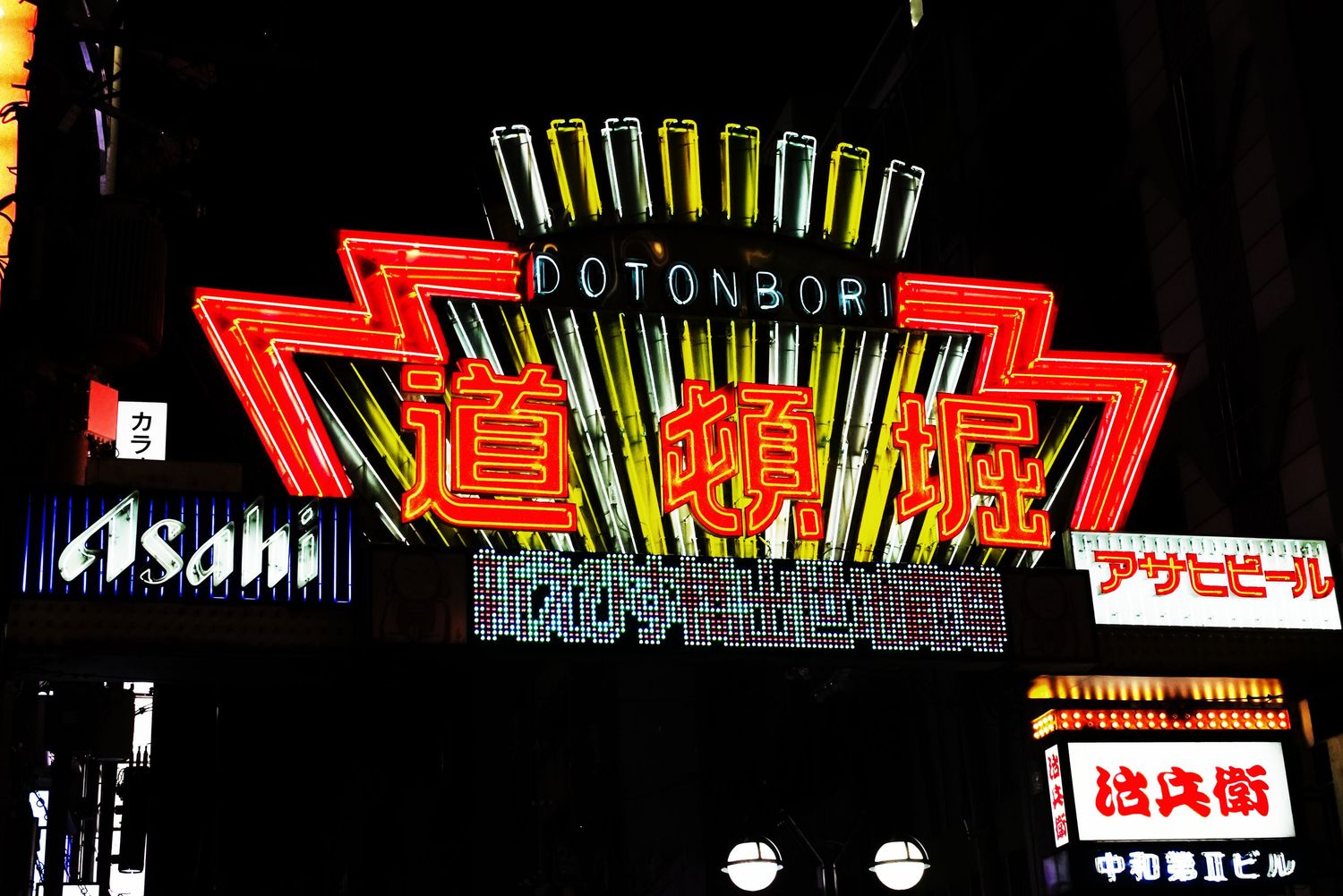 Bright neon signs illuminate the Dotonbori district in Osaka, Japan, featuring kanji characters and Asahi beer advertisements at night.