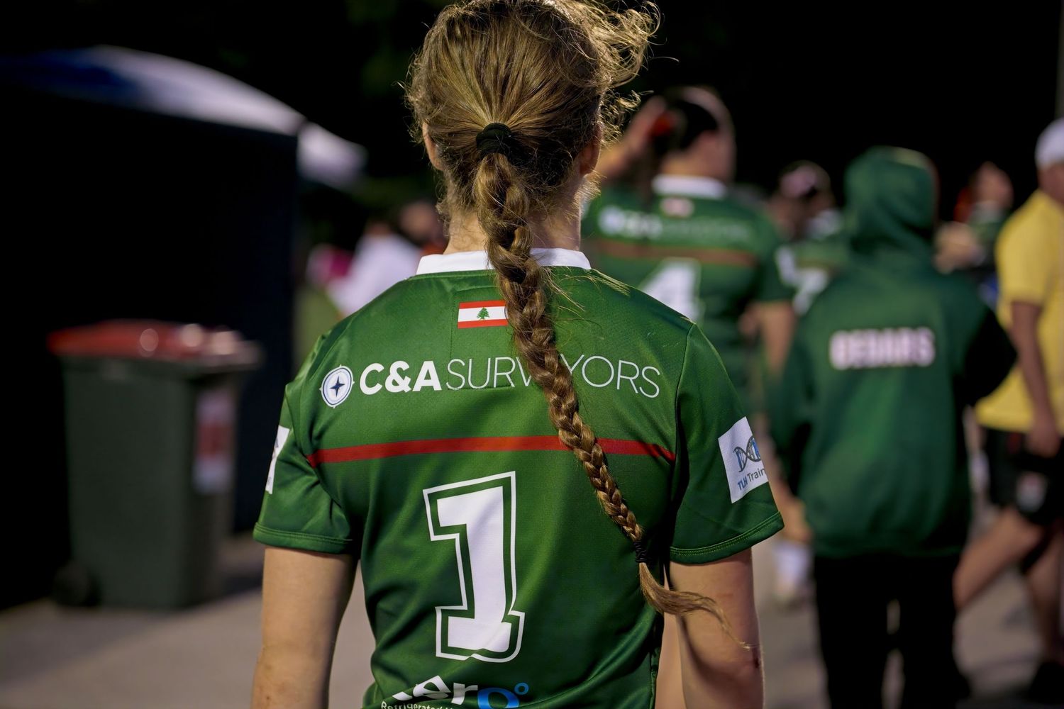 Girl in green C&A Surveyors jersey number 1 with braided hair stands at a sports event.
