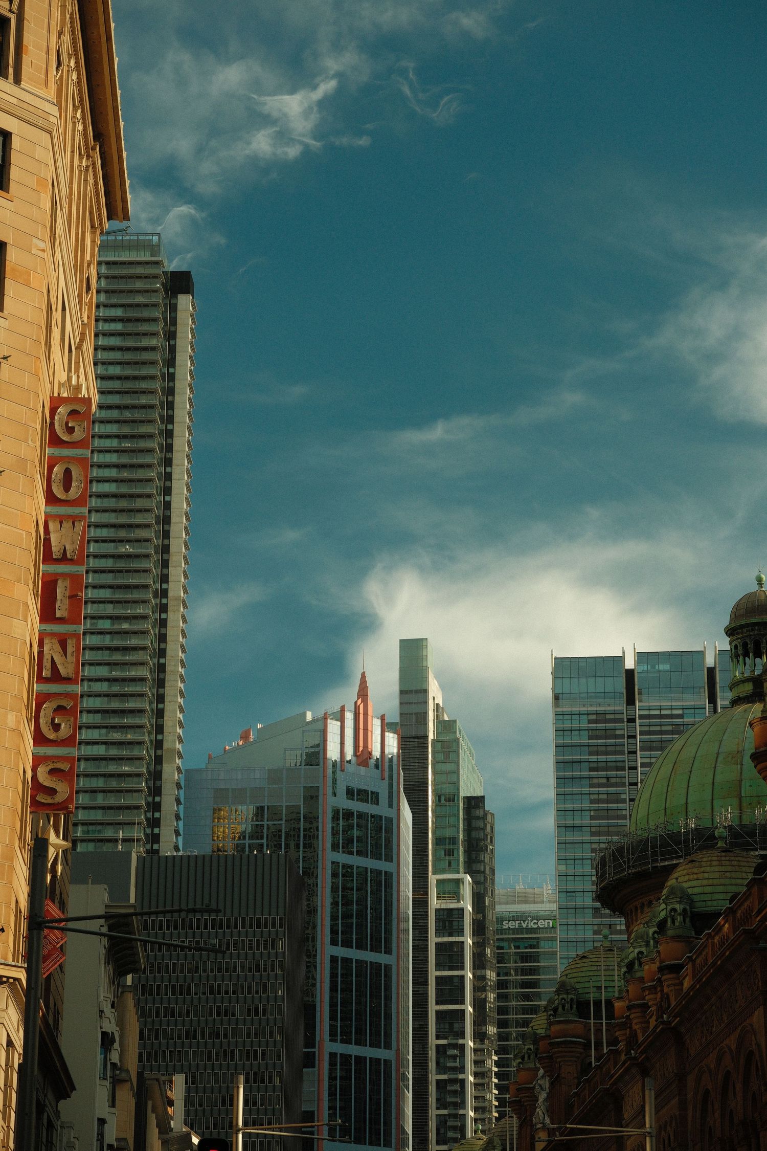 Sydney CBD skyline with Gowings building sign, modern glass skyscrapers, and historic dome under blue sky.