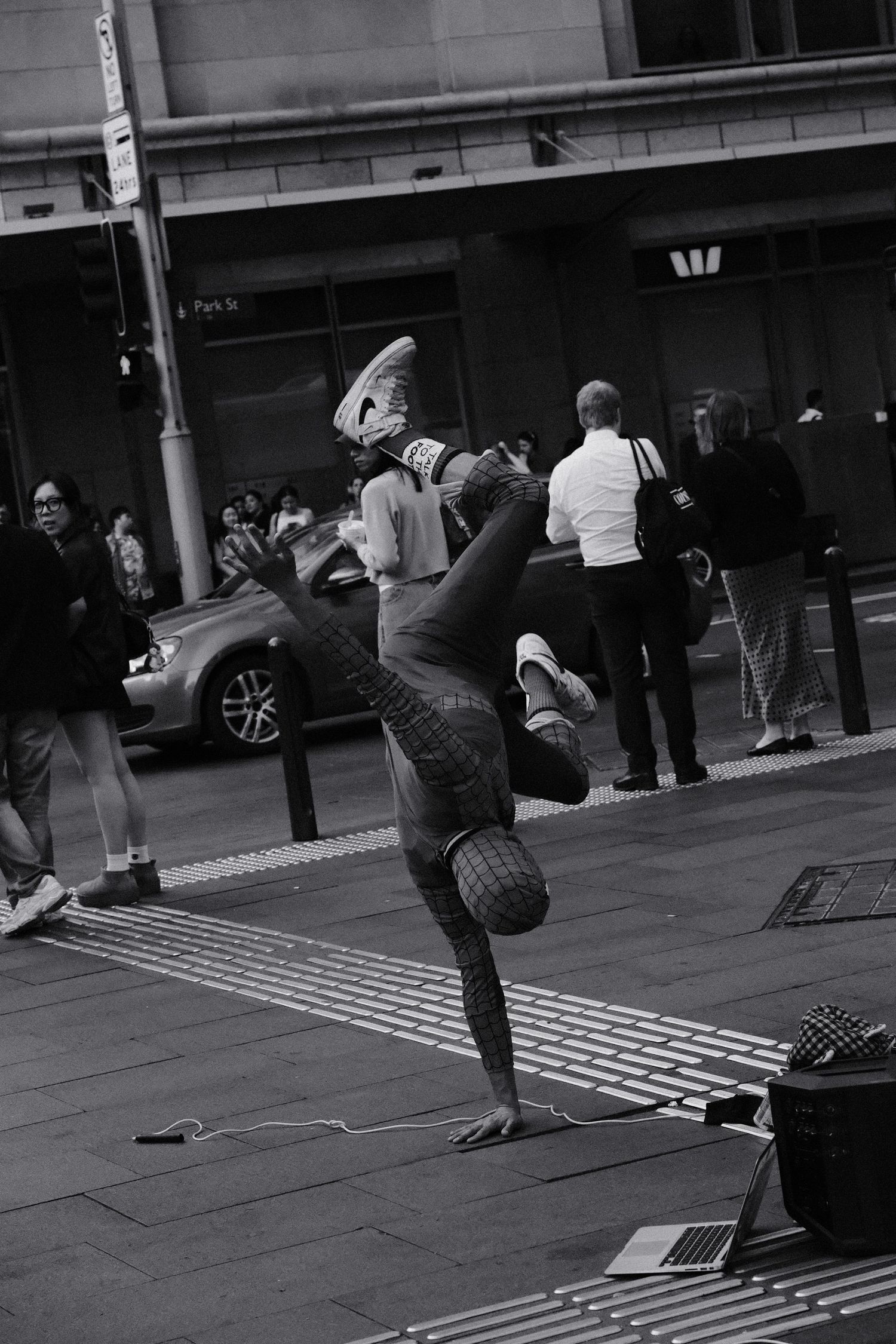 Street performer in Spiderman costume doing a one-handed handstand on a busy city sidewalk, black and white photo.