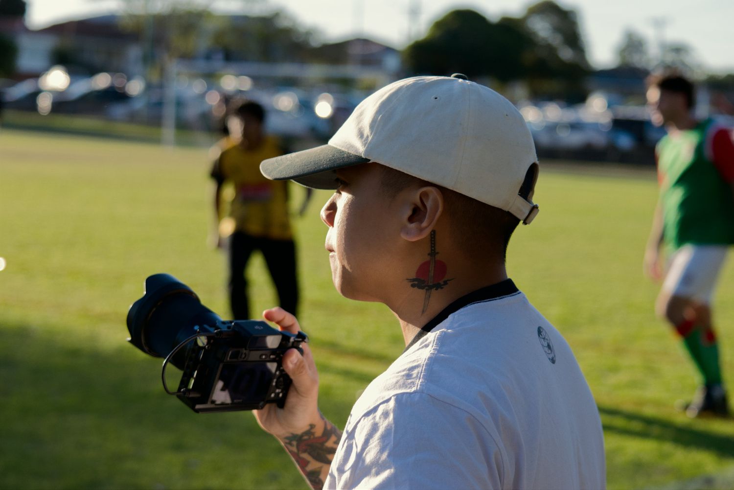 Person with ear tattoo filming a soccer match with a camera on a green field.