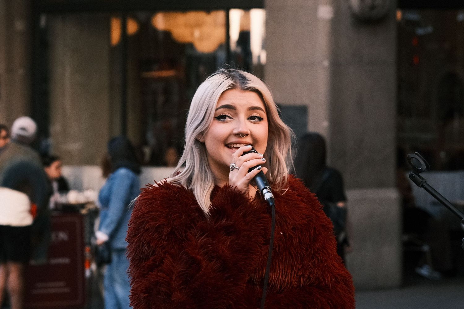 Young woman with blonde hair singing into a microphone outdoors, wearing a red fluffy coat.