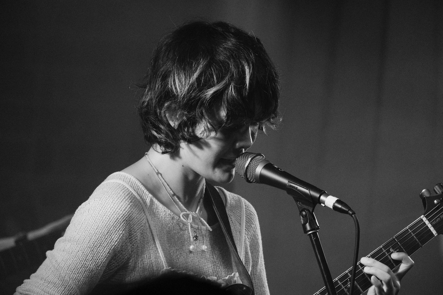 Black and white photo of a musician singing into a microphone while playing guitar on stage.