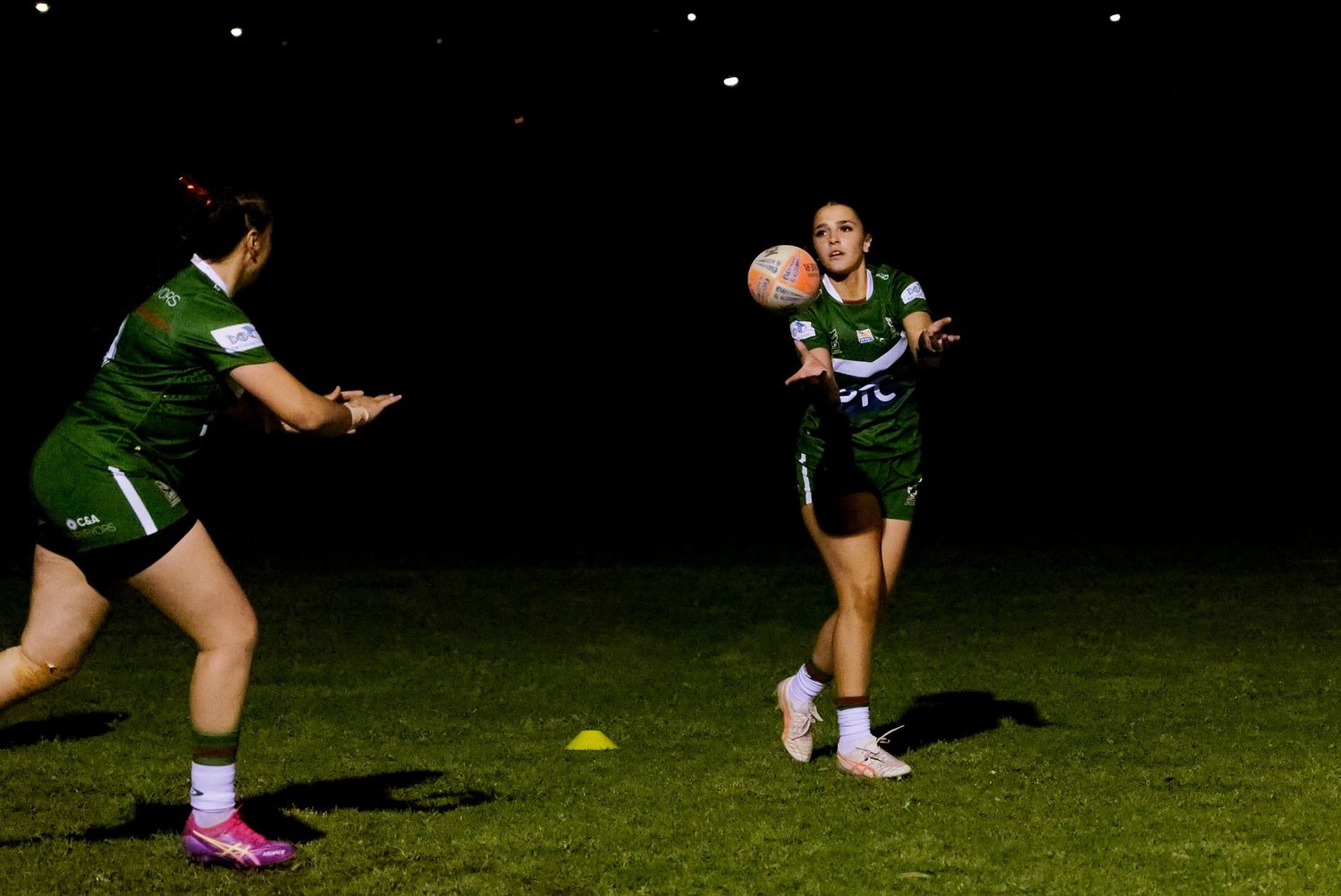Two women in green uniforms playing rugby under floodlights at night on a grass field.