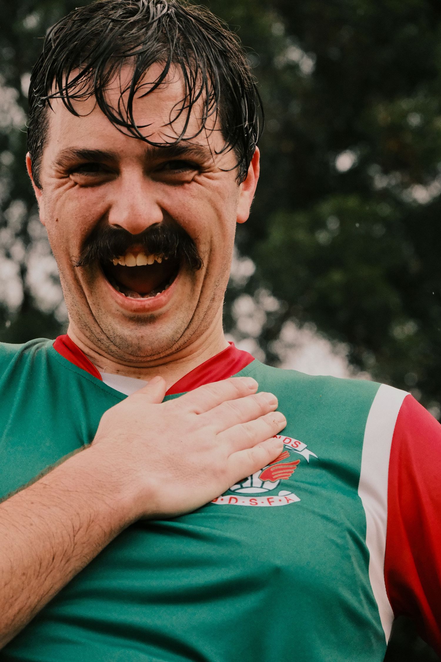 Excited man with mustache in green and red sports jersey, hand on chest, laughing outdoors.
