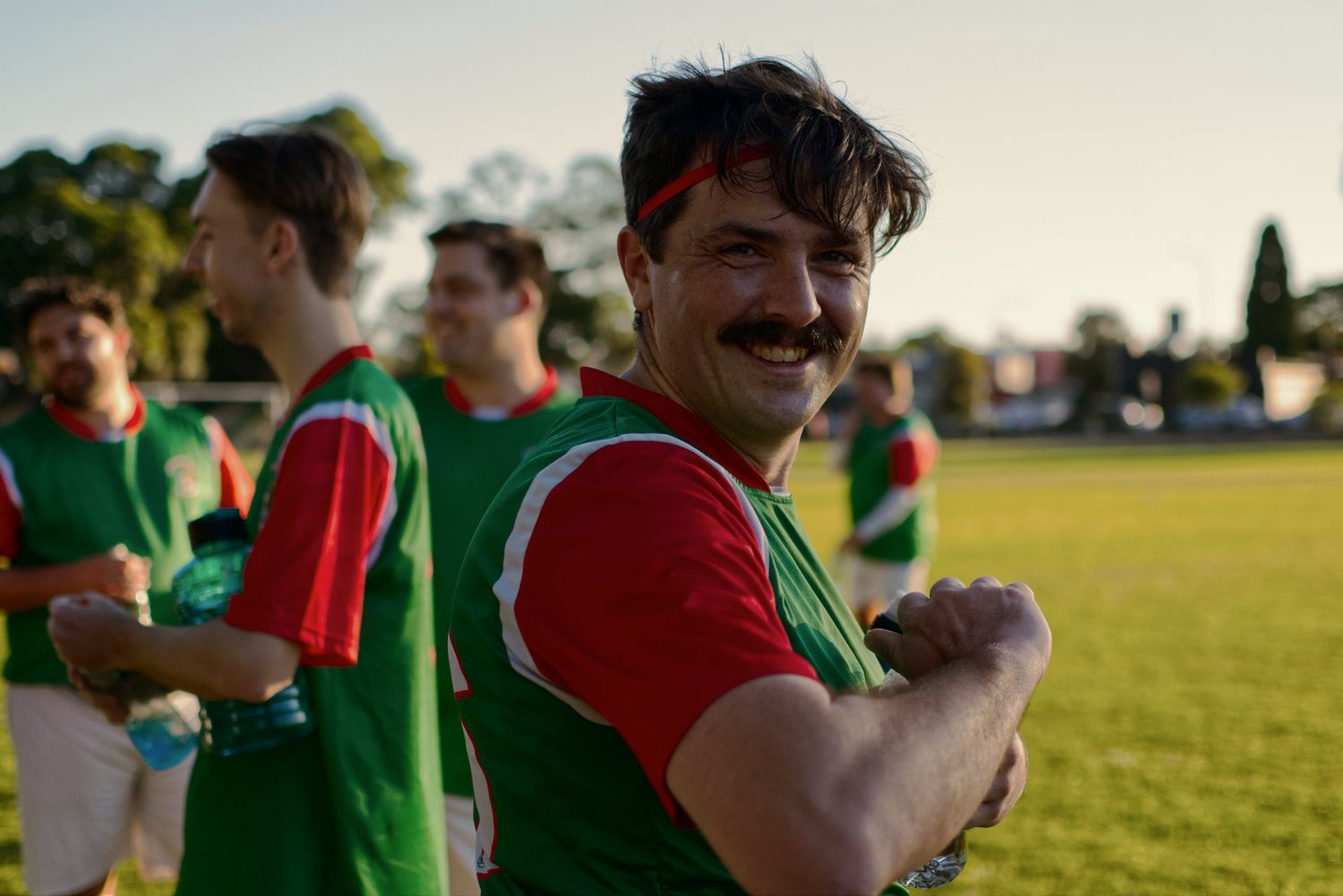 A smiling mustachioed soccer player in green and red jersey celebrates with teammates on field.