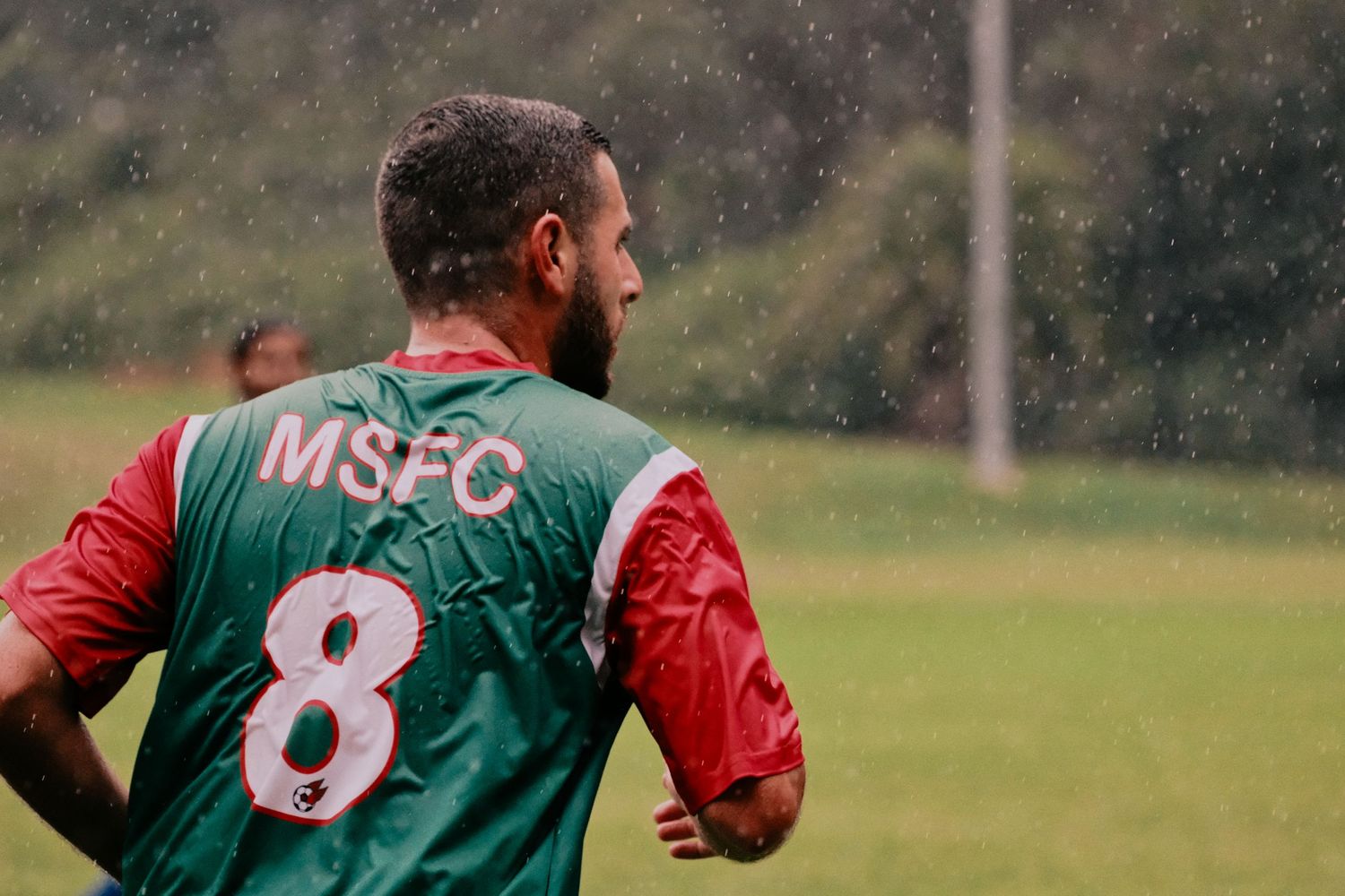 Bearded soccer player wearing MSFC number 8 green and red jersey playing in the rain on a grass field.