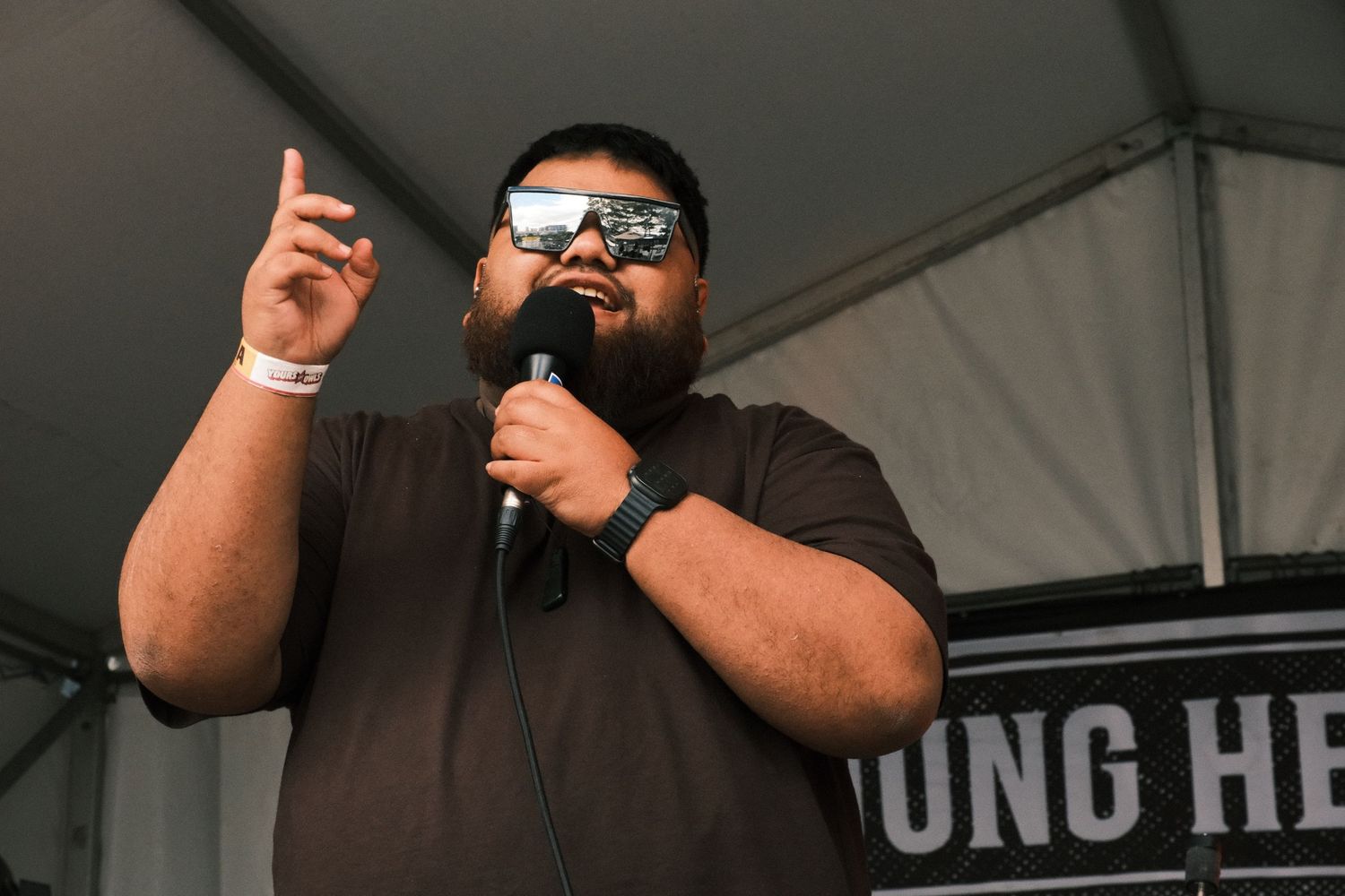 Man with beard and sunglasses speaking into microphone at outdoor event, pointing finger upward on stage.