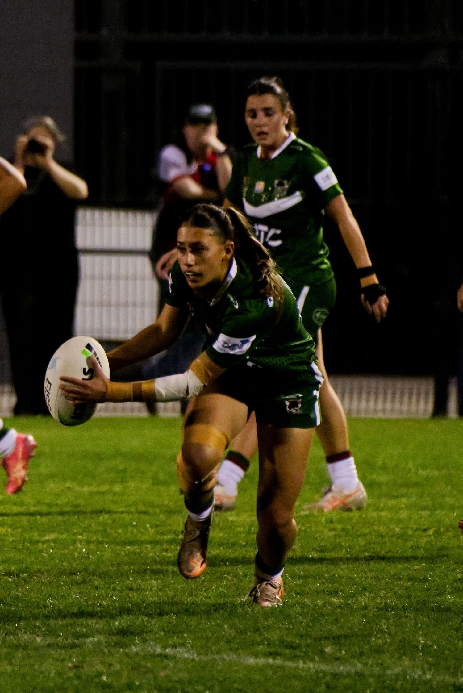 Female rugby league players in green uniforms compete during a night match, one diving to score a try.