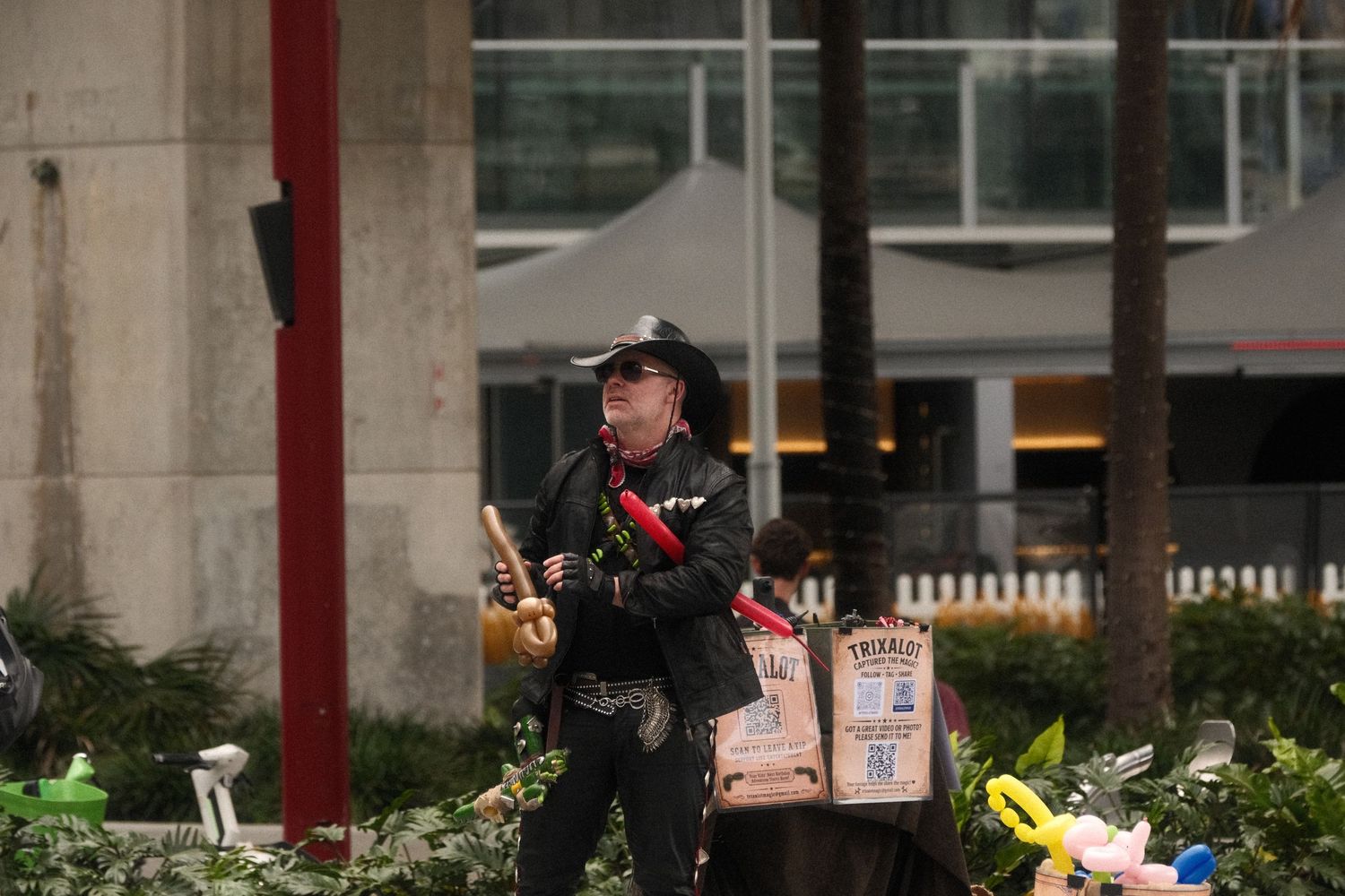 Street performer dressed as a cowboy juggling props outdoors near urban greenery and decorative displays.