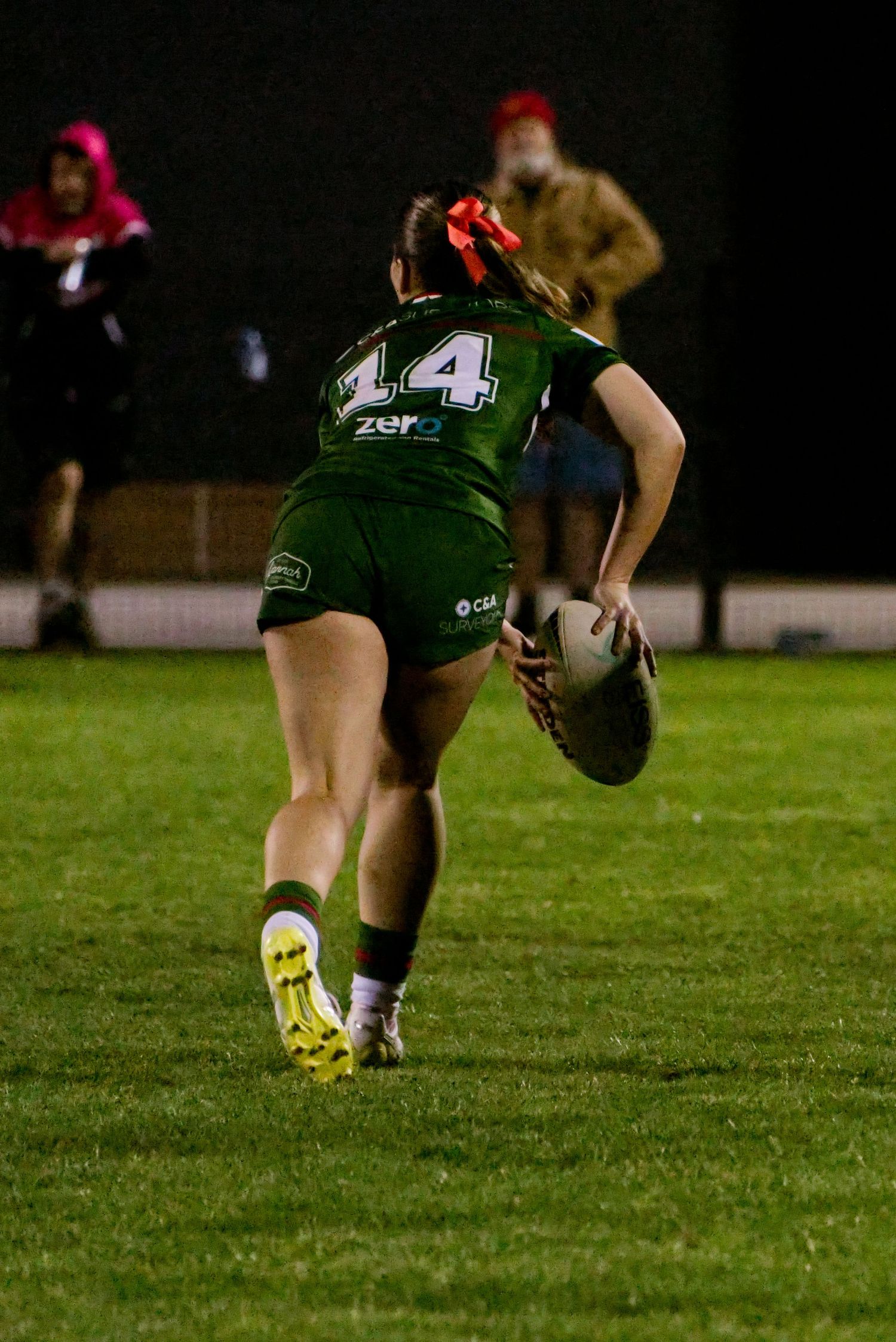 Female rugby league player #14 in green jersey runs with ball during night match on grass field.