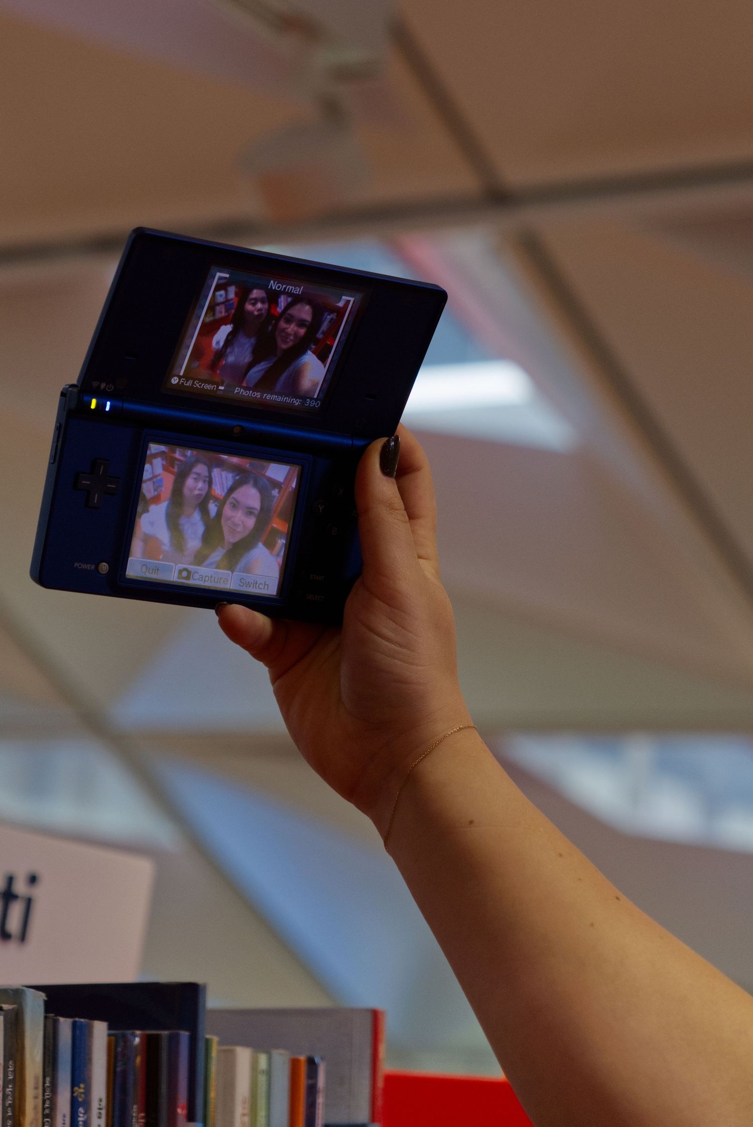 Person holding a Nintendo DSi handheld console displaying selfie photos on both screens in a library setting.
